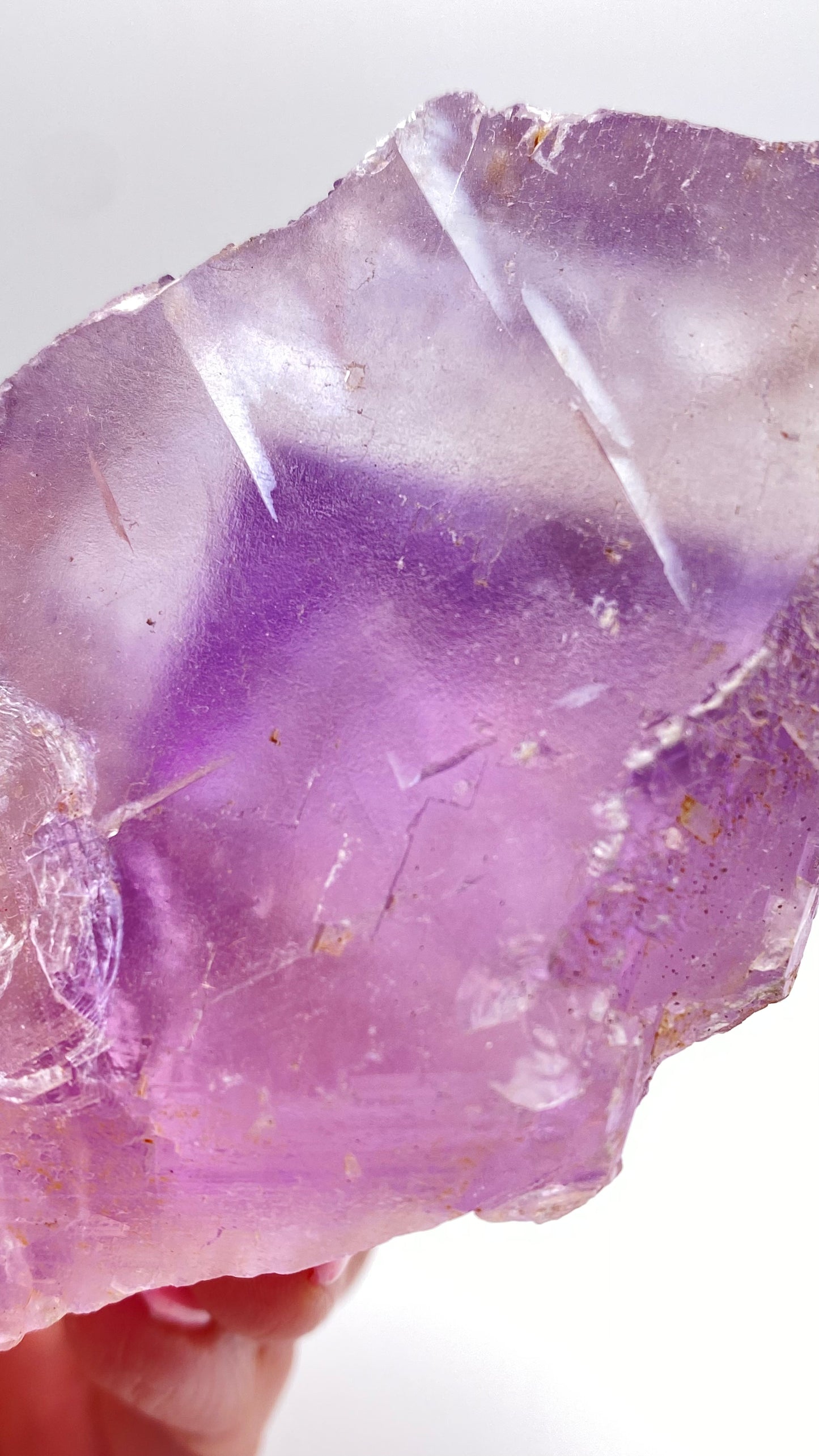 A close-up of a person's hand holding The Crystalary's Fluorite from Lead Hill, Cave-In-Rock, Hardin County, Illinois. This rough-textured, translucent crystal showcases an intricate pattern with shades of purple and visible cracks and imperfections against a plain white background.