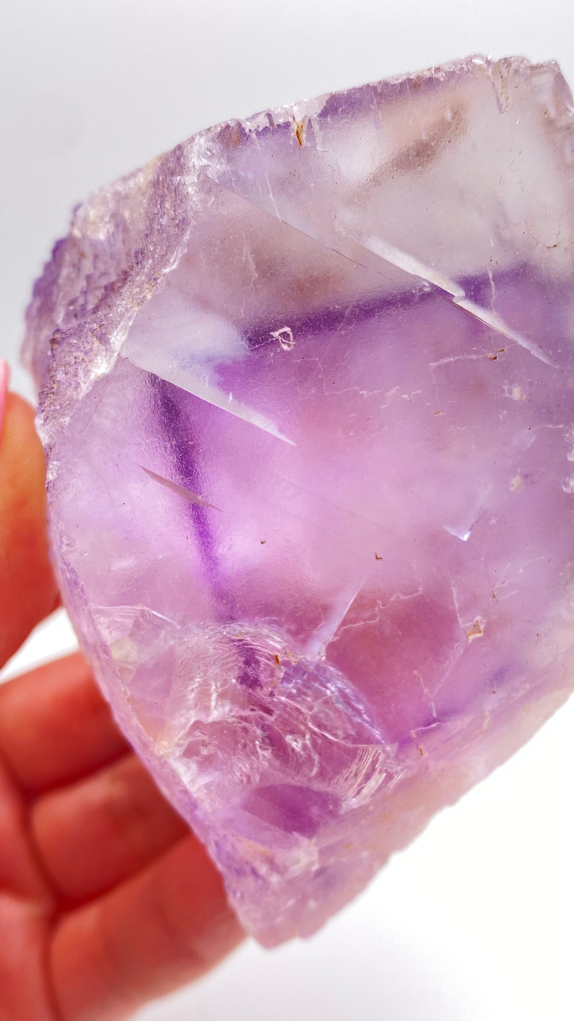 Close-up image of a hand holding a rough, translucent purple fluorite crystal with visible fractures and imperfections. The background is white, highlighting the vibrant color and texture of The Crystalary's Fluorite- Lead Hill, Cave-In-Rock, Hardin County, Illinois, USA specimen.