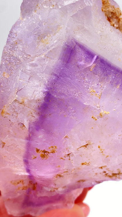 A close-up view of The Crystalary's Fluorite from Lead Hill, Cave-In-Rock, Hardin County, Illinois, USA showcases a stunning translucent purple crystal with a blend of light and dark purple hues. The specimen features irregular shapes and is adorned with several golden flecks scattered across its surface, adding textured details. A hand is partially visible holding the crystal.