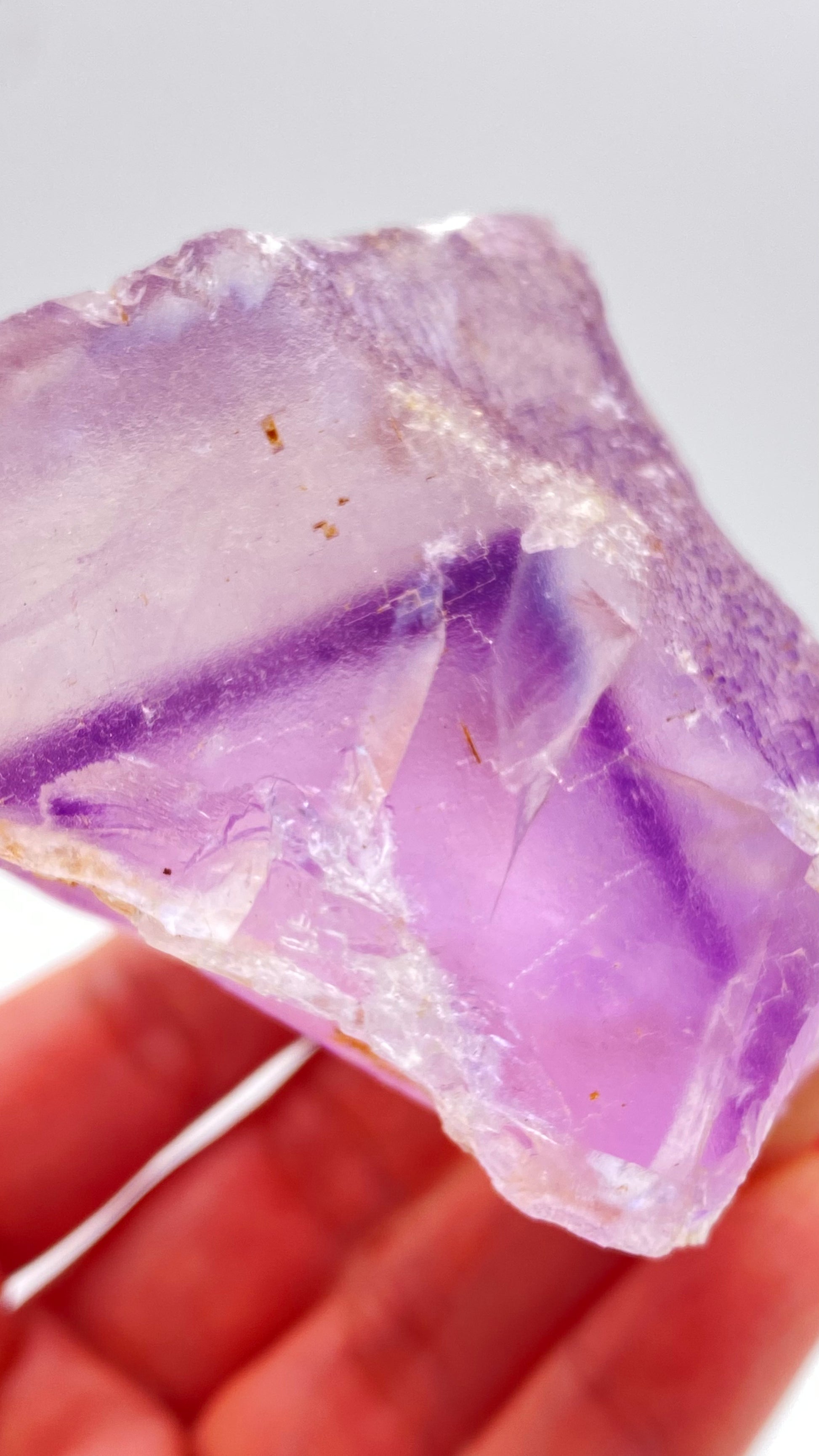 A close-up of a hand holding a translucent Fluorite specimen from The Crystalary, originating from Lead Hill, Cave-In-Rock, Hardin County, Illinois, USA. The specimen displays varying shades of purple and lavender and features a rough, uneven texture with small reflective surfaces catching the light. White background.

