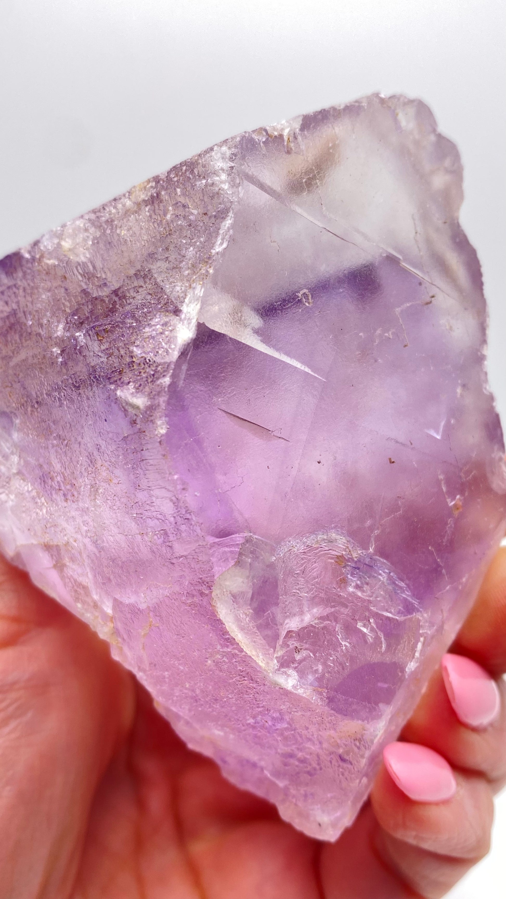 A close-up of a hand holding a translucent purple Fluorite crystal from Lead Hill, Cave-In-Rock, Hardin County, Illinois. The person has light skin and wears pink nail polish. The crystal's surface showcases intricate patterns and varying shades of purple, reminiscent of the specimens curated by The Crystalary. The background is plain white.