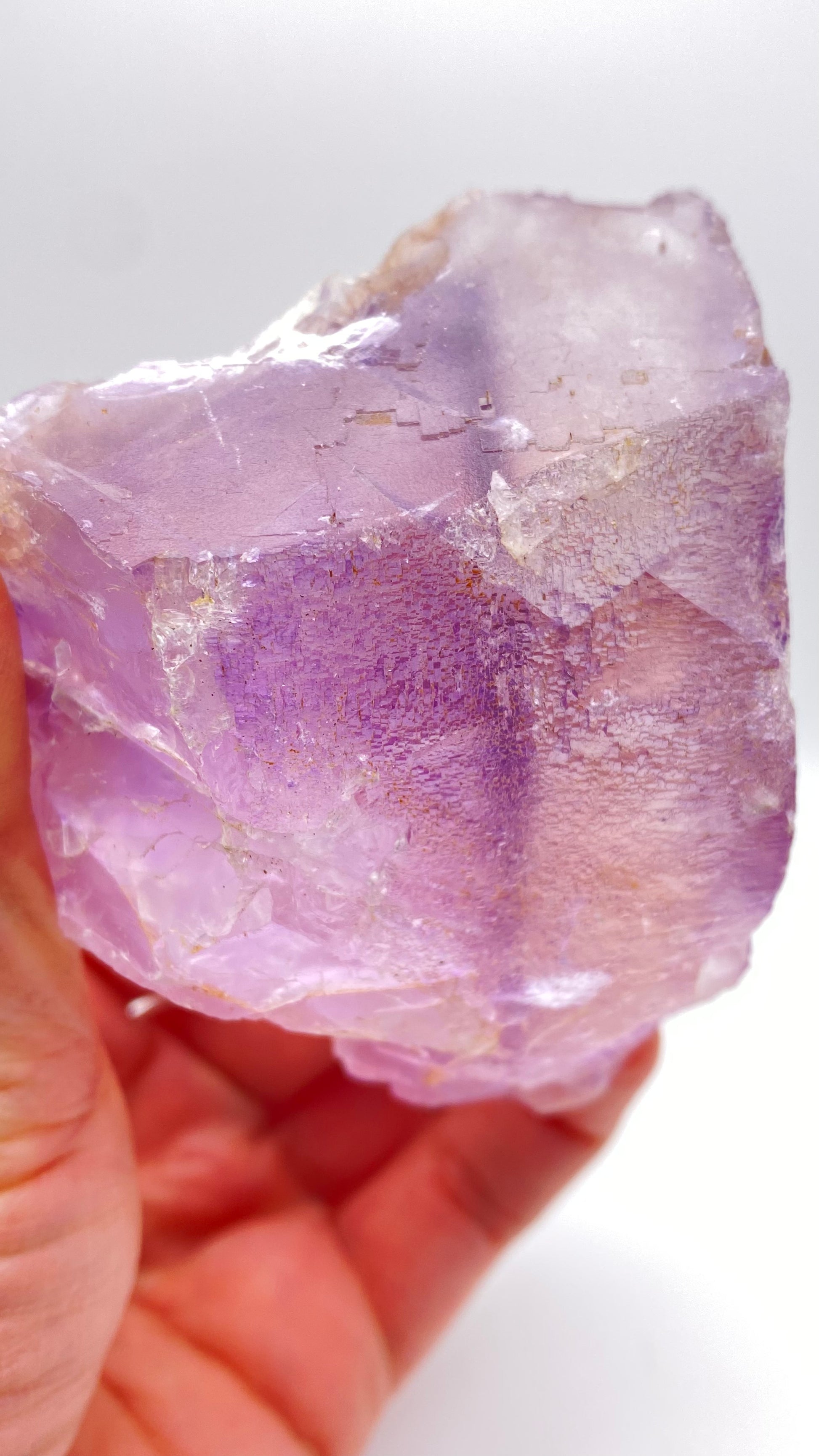 A hand holding a stunning Fluorite crystal from Lead Hill, Cave-In-Rock, Hardin County, Illinois by The Crystalary. This gem features rough and uneven surfaces with a captivating range of purple and lilac shades. It catches the light beautifully, revealing its semi-transparent and intricate interior structure—truly reminiscent of the treasured finds from Cave-In-Rock.