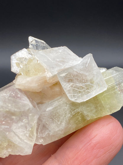 Close-up of a person's fingers holding a translucent, pale yellow-green Apophyllite crystal from The Crystalary's "Apophyllite, Stilbite" collection, sourced from Lonavala Quarry in the Pune District of Maharashtra, India. The crystal features irregular, angular facets and a slightly glossy surface against a gradient dark gray background.