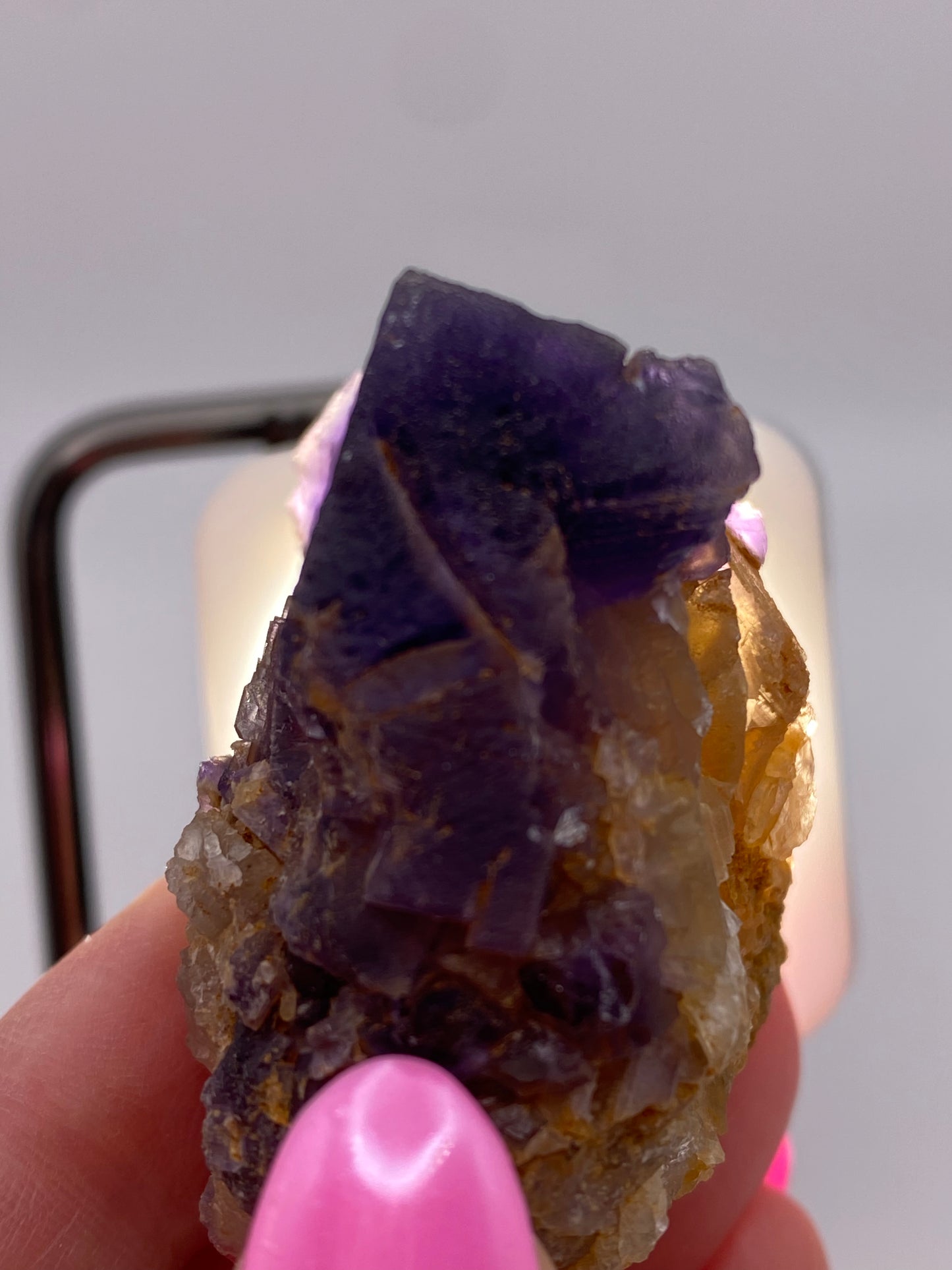 Close-up of a hand with pink-painted nails holding a raw, unpolished Fluorite from the Minerva Mine Number 1 in Hardin County, Illinois. The stone, from The Crystalary, is primarily dark purple with some translucent and yellowish-brown areas. A light source is visible in the background, illuminating the stone like focal cubes.