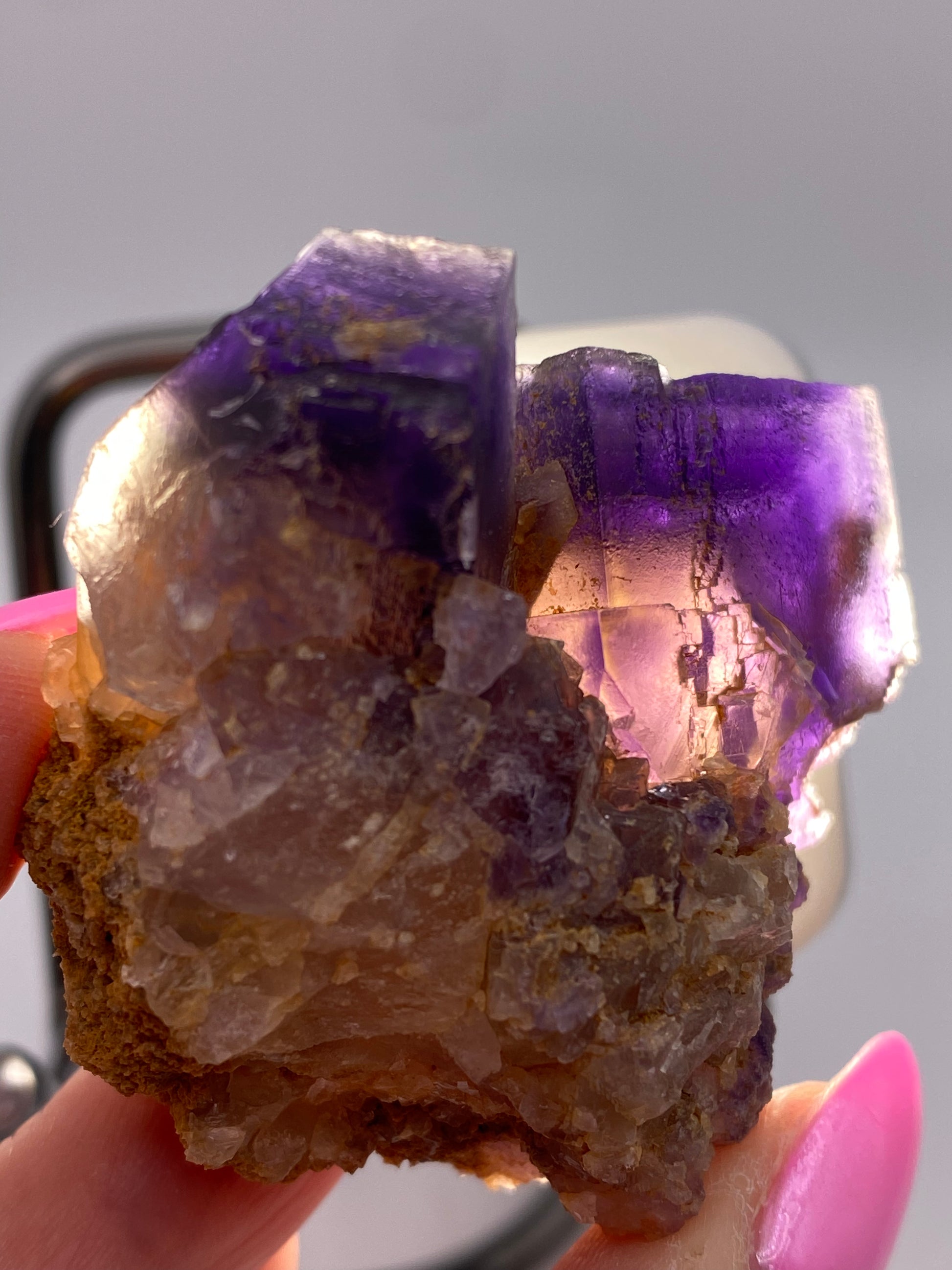 A close-up of a hand holding a rough mineral specimen from the Minerva Mine Number 1 in Hardin County, Illinois, displays the vibrant purple hues characteristic of The Crystalary's Fluorite. The mineral’s irregular surfaces and focal cubes gleam, while the nails of the hand are painted pink against a blurry background.