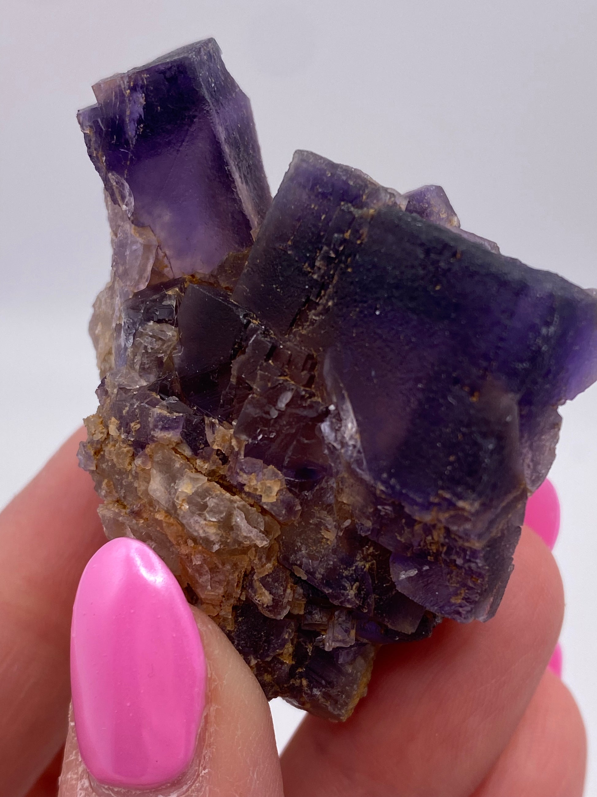 Close-up of a hand holding a Fluorite mineral from The Crystalary collection at Minerva Mine Number 1 in Hardin County, Illinois. The dark purple fluorite has rough, angular surfaces and is partially translucent, revealing internal facets. The hand’s neatly manicured nails are painted with bright pink polish, providing a striking contrast to the mineral's deep hues.