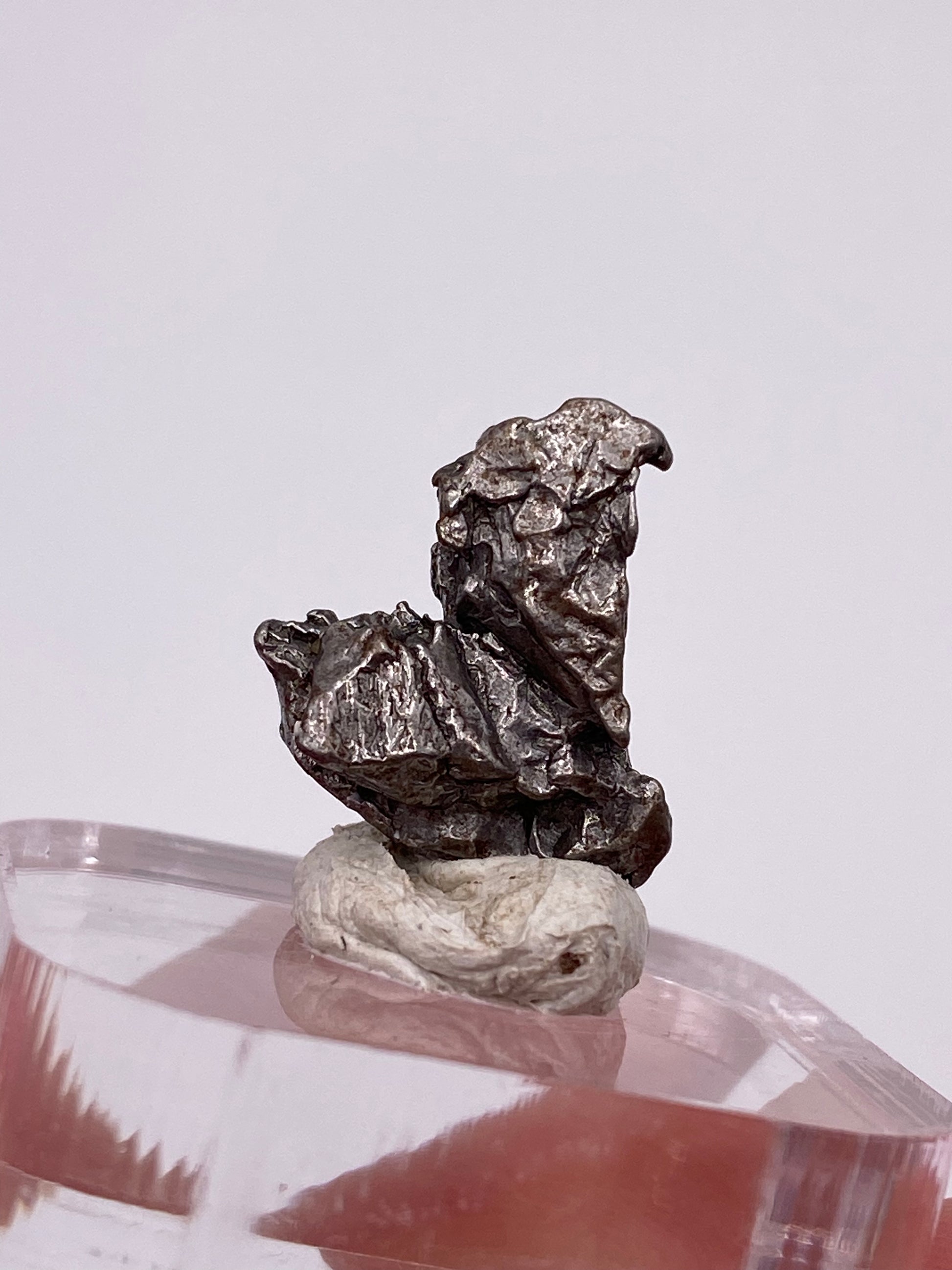 A close-up image of a silvery-gray metallic mineral specimen, the Campo del Cielo Meteorite from Gancedo, Doce de Octubre Department, Chaco Province, Argentina. This product by The Crystalary features a rough, jagged surface and is mounted on a white putty-like substance atop a clear acrylic base. The plain, light-colored background highlights the texture and color of this remarkable meteorite.