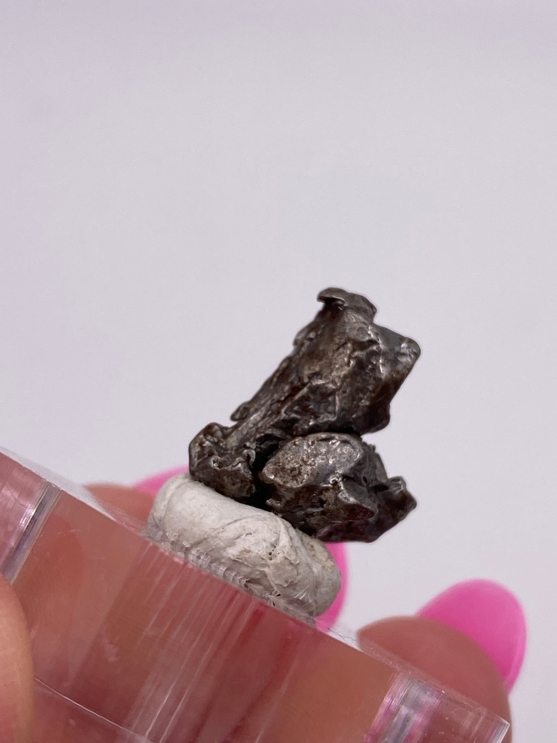 A close-up of a hand holding a small, rough chunk of the Campo del Cielo Meteorite- Gancedo from The Crystalary atop a white substance. The nugget balances on a clear cylindrical platform, highlighting its iron meteorite structure. The background is blurred, emphasizing the Argentine metallic treasure and the person's pink-painted nails.