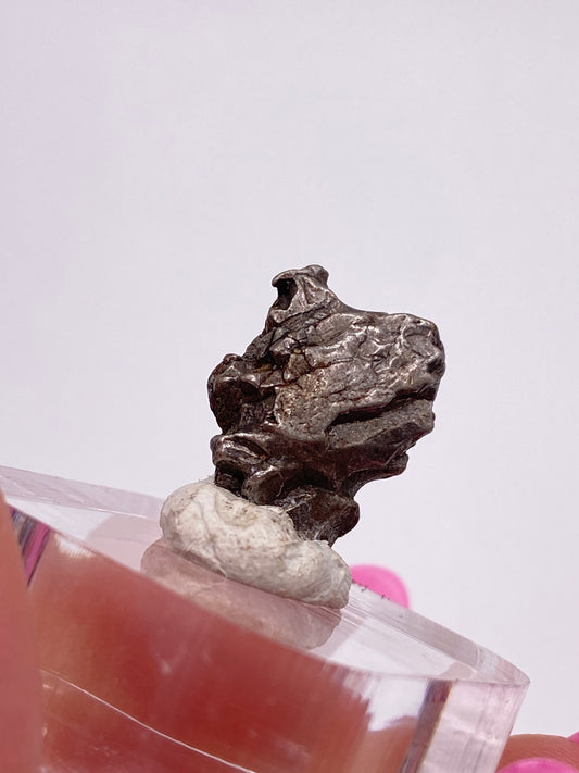 A small metallic Campo del Cielo Meteorite fragment from Gancedo in the Doce de Octubre Department of Argentina's Chaco Province, displayed on a clear cube stand with a white tack at the base, set against a plain white background, by The Crystalary.