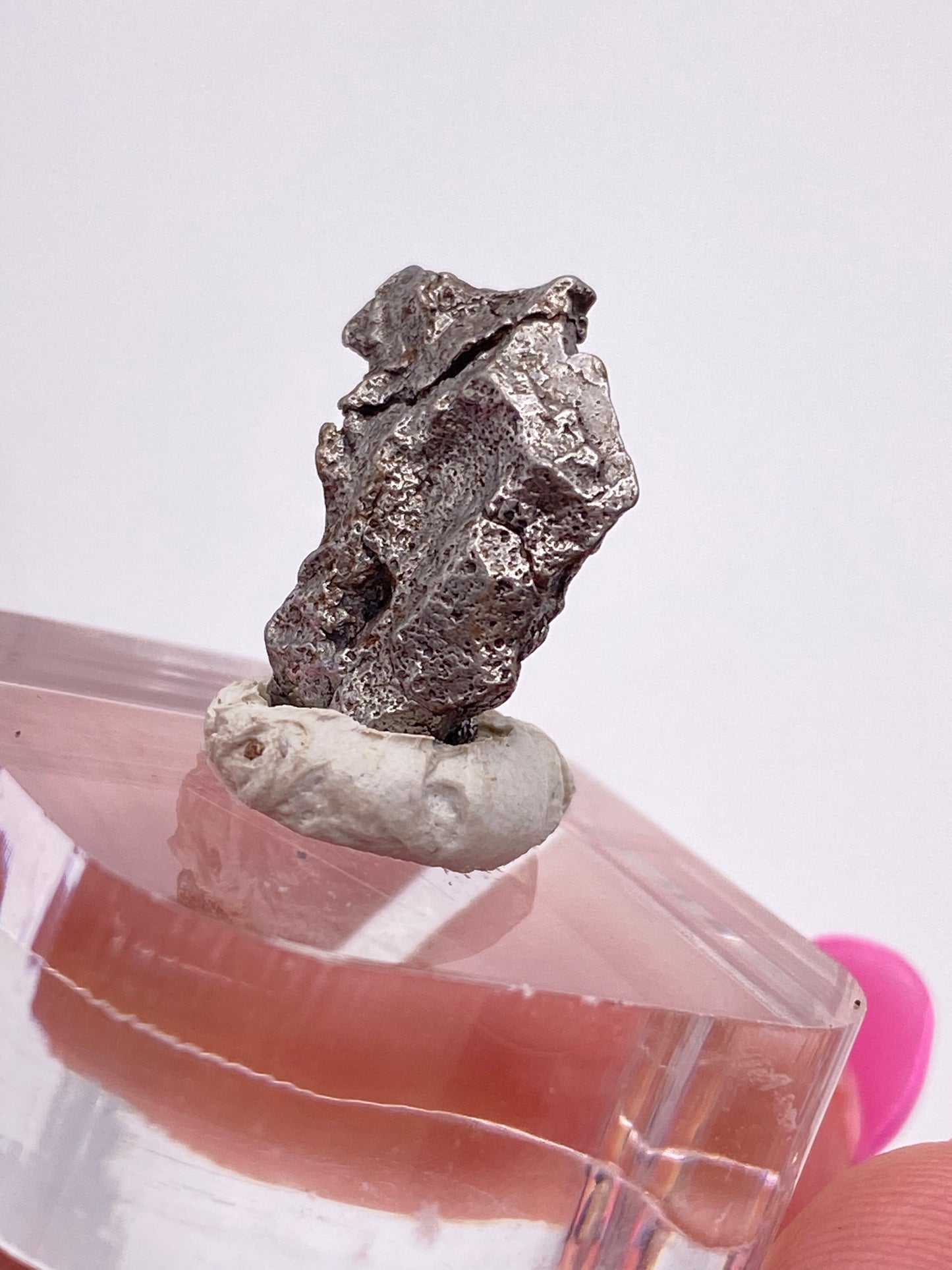 A close-up image of a piece of silver metallic rock, the Campo del Cielo Meteorite from Gancedo in the Doce de Octubre Department, Chaco Province, Argentina, perched on a small white clay base atop a clear geometric stand. The background is out of focus, which emphasizes the metallic texture and contrast. This remarkable specimen is part of The Crystalary collection.