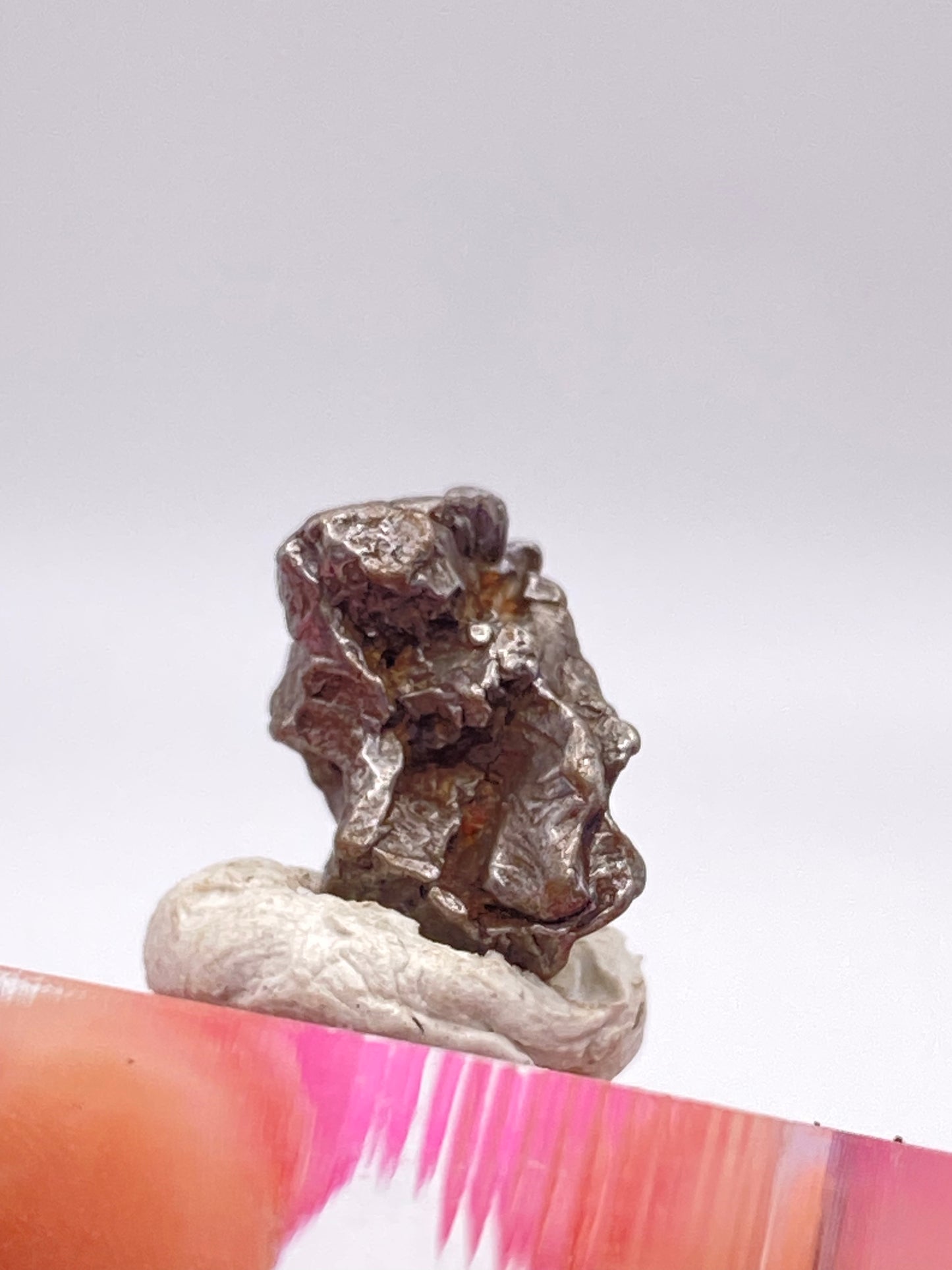 Close-up of a small piece of metallic rock from the Campo del Cielo Meteorite - Gancedo, Doce de Octubre Department, Chaco Province, Argentina by The Crystalary, showcased against a white background. The irregularly shaped object has a dark, rough, and shiny surface and is positioned on a pink and white base.