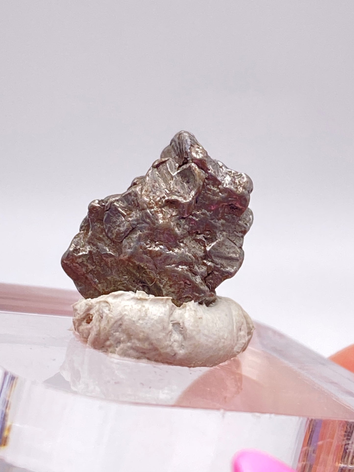 A rough, metallic rock, identified as the Campo del Cielo Meteorite- Gancedo from Doce de Octubre Department, Chaco Province, Argentina by The Crystalary, is displayed on a small white clay pedestal. The uneven surface of this iron meteorite has a dark, shiny appearance against a plain, light-colored background. The setup rests on a clear textured platform.