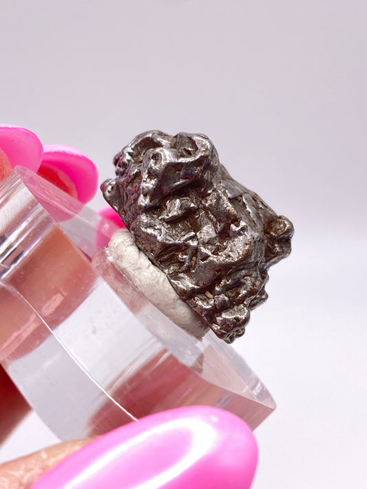 A hand with pink-painted nails holds a small, rough metallic nugget, identified as The Crystalary's Campo del Cielo Meteorite from Gancedo, Doce de Octubre Department in Chaco Province, Argentina, mounted on a clear plastic stand against a plain white background.