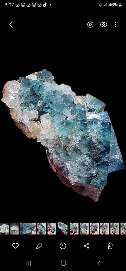 A detailed close-up photograph showcases a large, vibrant Fluorite- Supernova Pocket crystal cluster from the Diana Marie Mine, Weardale, Co. Durham, England. This striking piece by The Crystalary features various blue and green facets intermingled with smaller white quartz crystals. Set against a solid black background, the colors and textures of these minerals are highlighted beautifully.