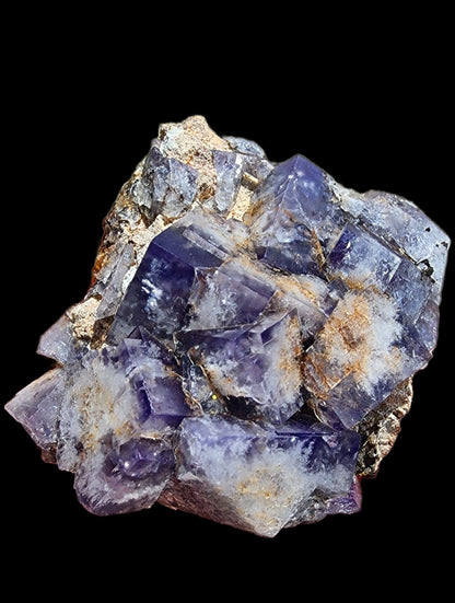 The Fluorite-Milky Way Pocket from The Crystalary, sourced from the Diana Maria Mine in Weardale, Co Durham, UK, features a cluster of deep purple crystal formations accented with gold and white mineral deposits set against a black backdrop. These crystals resemble the geometric structure and sharp edges characteristic of fluorite and display varying levels of opacity, evoking images of tiny Milky Way Pockets.