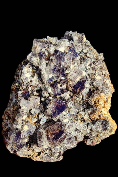 A cluster of rough, purple, and white Fluorite crystals from the Smoky Rabbit Pocket at the Diana Maria Mine rests on a dark background. The jagged surface varies in size and form, reflecting light at different angles, showcasing the enchanting allure of Weardale's treasures. From The Crystalary collection (sku3015).