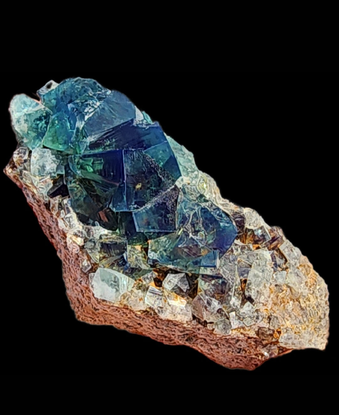 A vibrant mineral specimen from the renowned Greedy Hog pocket at the Diana Maria Mine, Co Durham, UK, features deep blue and green crystals with a rough, earthy base. The Fluorite's large central crystals are transparent, displaying intricate patterns and facets against a black background. This stunning piece is brought to you by The Crystalary (Product SKU 3071).