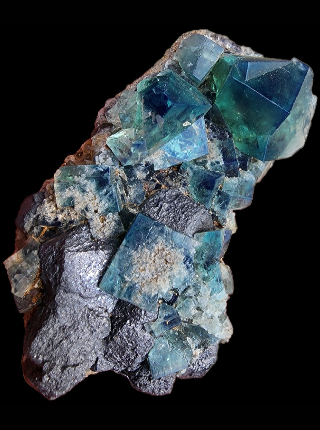 A striking arrangement of fluorite crystals in vibrant blue and green hues, sourced from the renowned Greedy Hog Pocket at the Diana Maria Mine in Co Durham, UK (sku 3062), rests on a dark gray rocky base. The crystals exhibit sharp, angular formations with colors transitioning from deep oceanic tones to lighter, translucent shades, all beautifully highlighted against a black background. This exquisite piece is offered by The Crystalary.