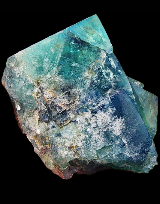 A close-up of a vibrant, multi-faceted Fluorite crystal from the Diana Maria Mine in Weardale, Co Durham, UK. The mineral, exhibiting gem-like qualities with shades of blues and greens, has a jagged, textured surface with areas of translucency and embedded impurities. It is set against a stark black background. Presenting: Fluorite- Truffle Pig Pocket by The Crystalary.