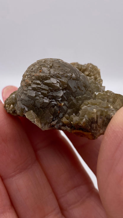 A close-up of a hand holding a rough, greenish-gray mineral specimen with a textured, crystal-like surface. The background is a plain white surface, which helps to highlight the details of the Adamite from Ojuela Mine in Mapimí, Durango, Mexico by The Crystalary.