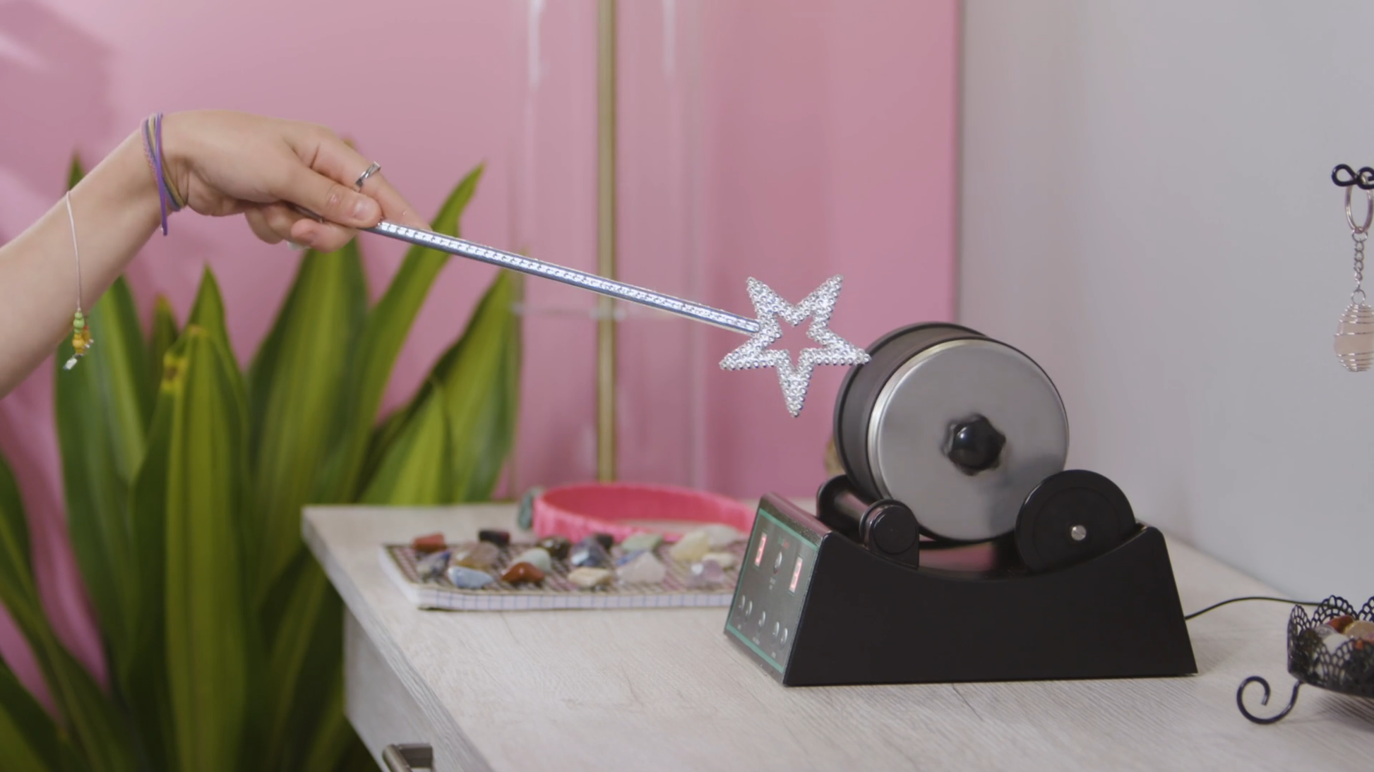 A person wields a sparkling star wand beside the Dan&Darci Advanced Rock Tumbler on a table. Neatly organized stones and a pink container adorn the surface. In the background, pink walls and green plant leaves complement the scene of rock tumbling magic.