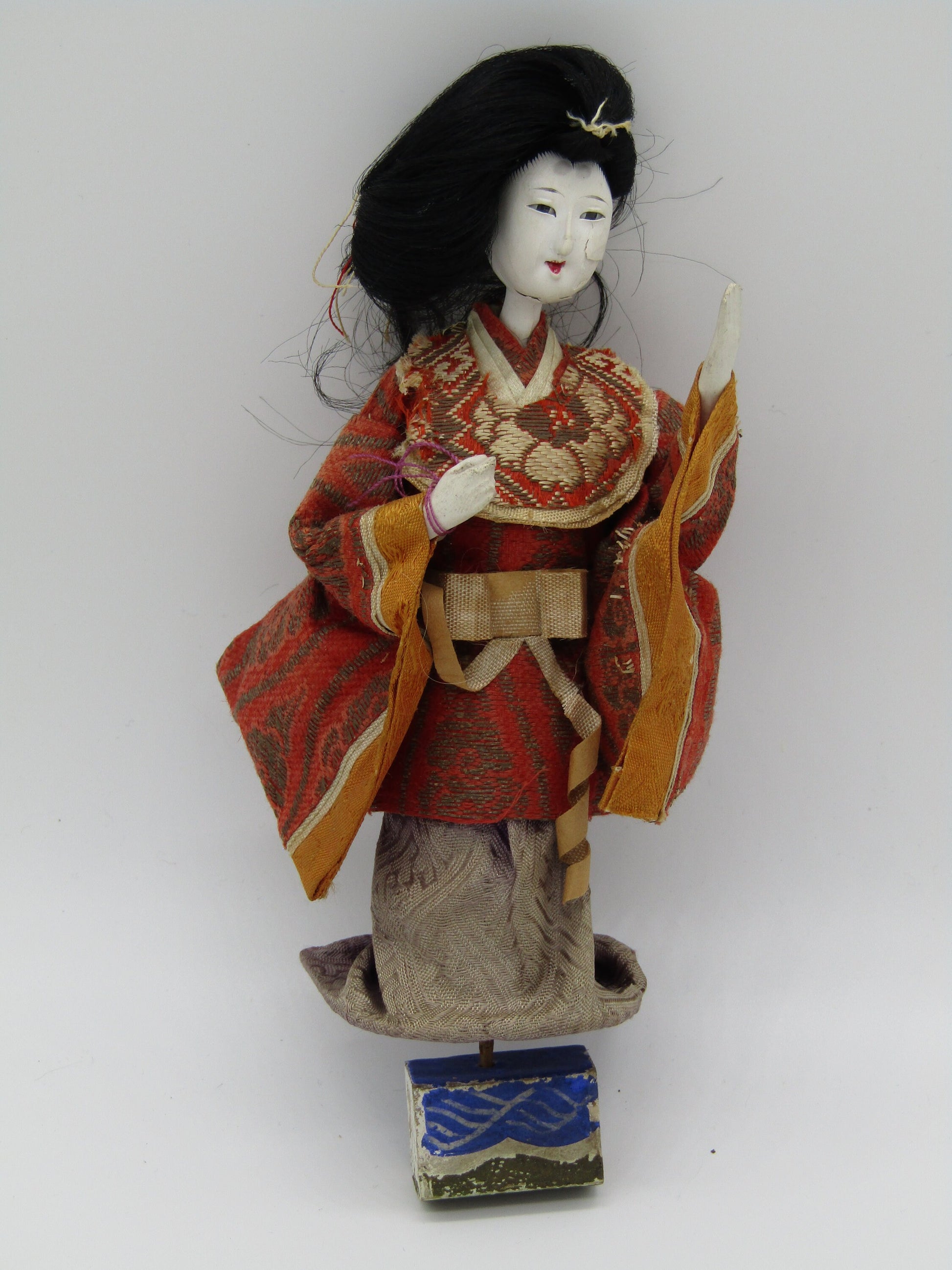 The Crystalary's Antique Japanese Ningyo Doll, an Asian doll from the early to mid-20th century, stands at 7 inches tall. This vintage Ningyo doll features a red and orange patterned kimono with beige detailing and a cream-colored obi belt. It has long black hair, a painted white face with serene features, and stands on a small blue and white platform.