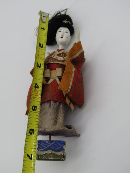 An antique Japanese Ningyo doll from The Crystalary, named "Antique Japanese Ningyo Doll Asian Doll 7" Early - Mid 20th Century Woman Gofun," stands upright with detailed clothing beside a yellow measuring tape, indicating its height is about 7 inches. The vintage doll wears a red and orange kimono and has black hair styled up.