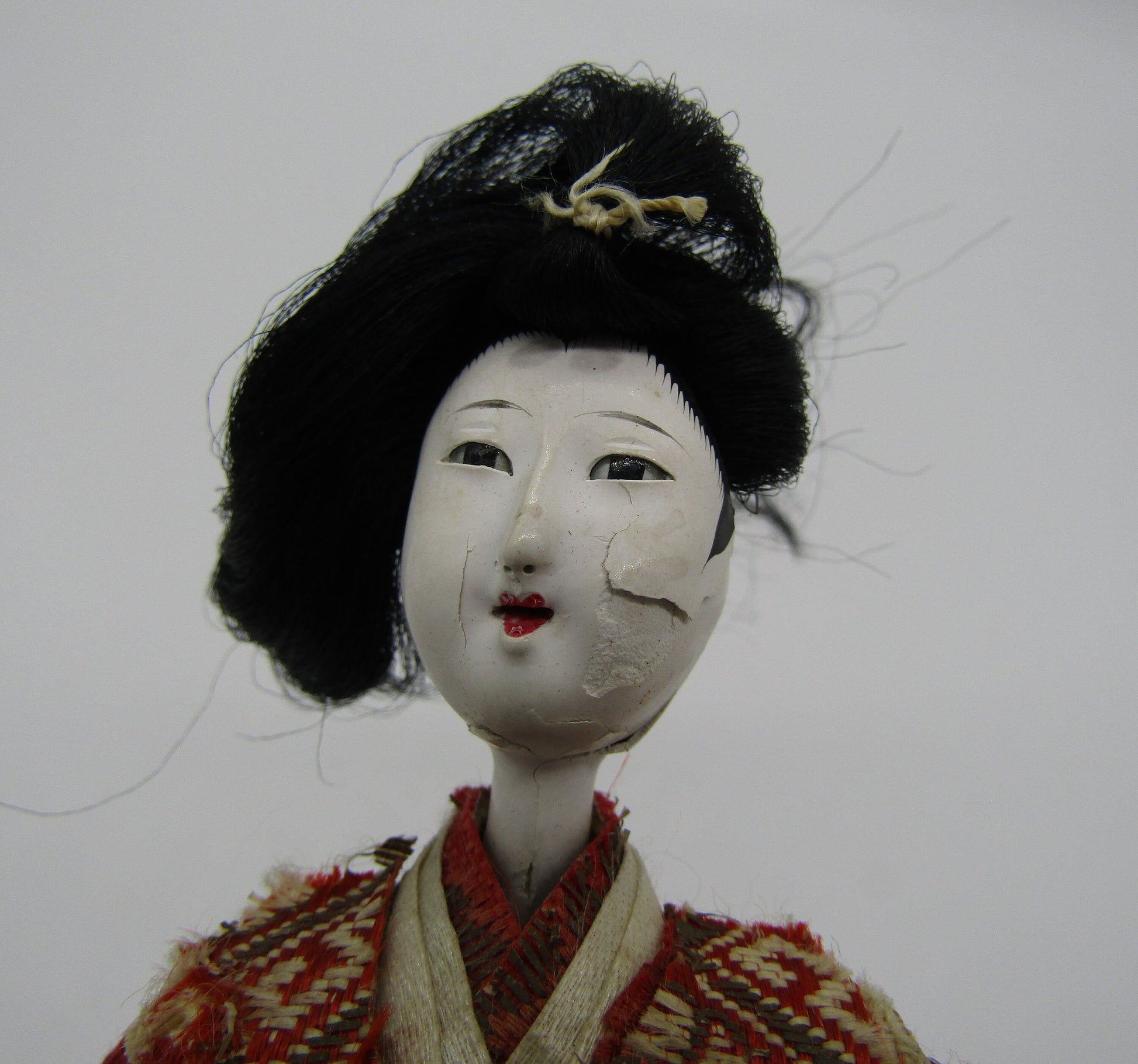 A close-up of The Crystalary's Antique Japanese Ningyo Doll showcases its intricately painted white face, despite some cracking and peeling. This 7" tall early to mid-20th century doll features black hair styled in an elegant updo, adorned with a delicate hairpin, and is dressed in a beautifully crafted red and beige kimono. The light-colored background further highlights the vintage beauty of this Gofun woman doll.