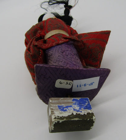 An Antique Japanese Ningyo Doll, crafted by The Crystalary and featuring a 7" Early - Mid 20th Century Woman Gofun design, is dressed in a red and purple traditional garment. The vintage doll is lying on its side with a partially worn square base at the bottom. It has a label attached with the numbers '15.9' and another larger label reading 'JP-9-11.' The background is a plain, neutral surface.