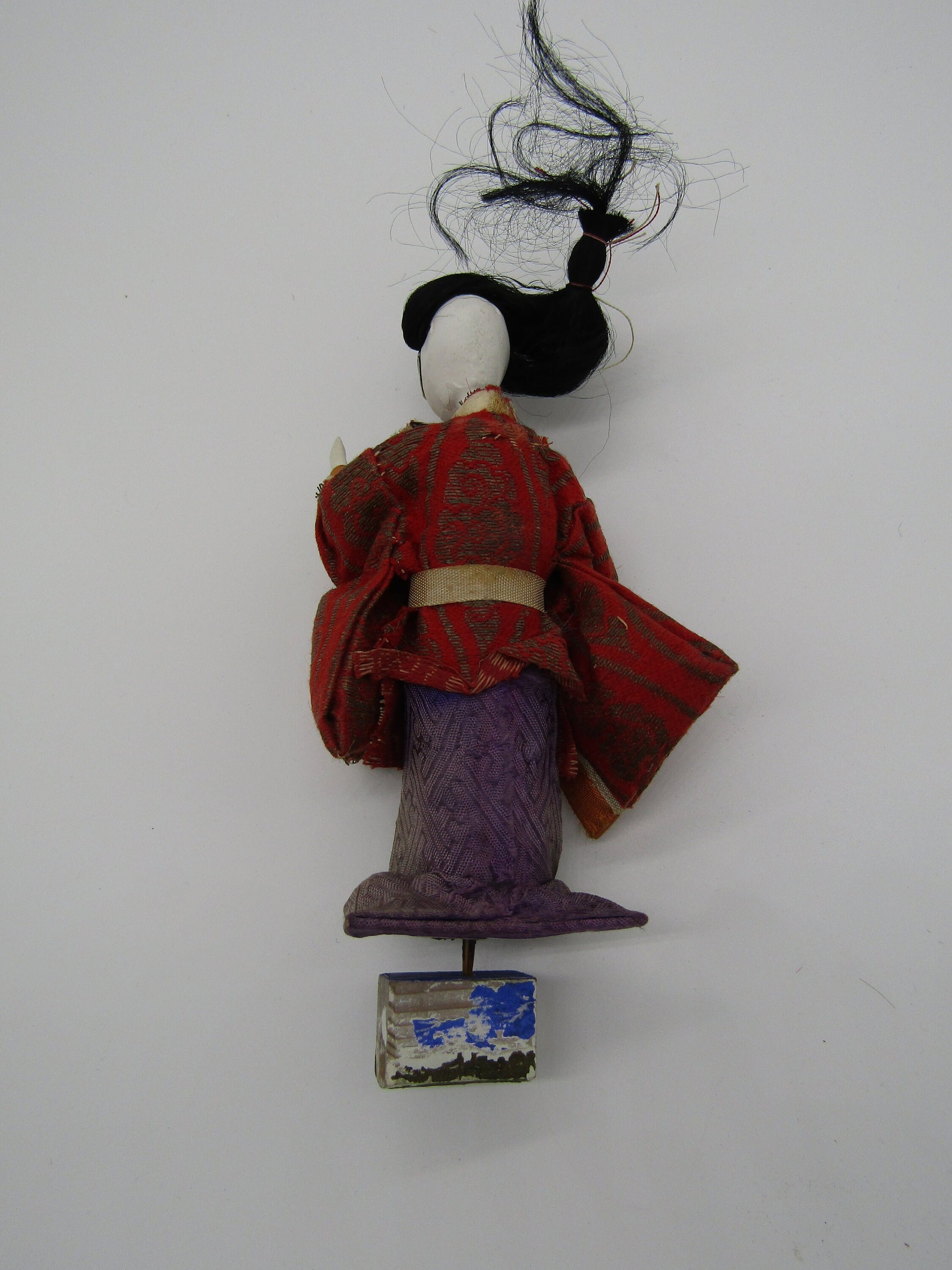 A photo of the Antique Japanese Ningyo Doll by The Crystalary. This 7" Asian doll, dating from the early to mid-20th century, is dressed in a vintage red kimono adorned with gold patterns and has a purple skirt. The figure of a Japanese woman features black hair styled up and stands on a small pedestal with white and blue painted designs. The face is an unadorned white surface without features.