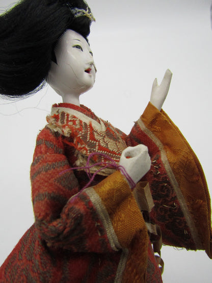 A close-up of *The Crystalary's* Antique Japanese Ningyo Doll, an exquisite 7" figure from the early to mid-20th century. The woman is dressed in an intricate orange and gold kimono, featuring a pale gofun face with delicate features and black, elaborately styled hair. One arm is raised, creating a graceful pose against a plain, light background.