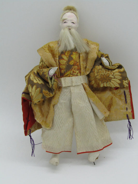 Product Description: The Antique Japanese Ningyo Doll Man from The Crystalary showcases a traditional ensemble, adorned in an ornate, gold-patterned kimono with extended sleeves. This meticulously crafted doll features a long white beard and hair styled in a topknot. The exquisite attire is decorated with floral and leaf designs and finished with purple tassels at the ends of the sleeves.