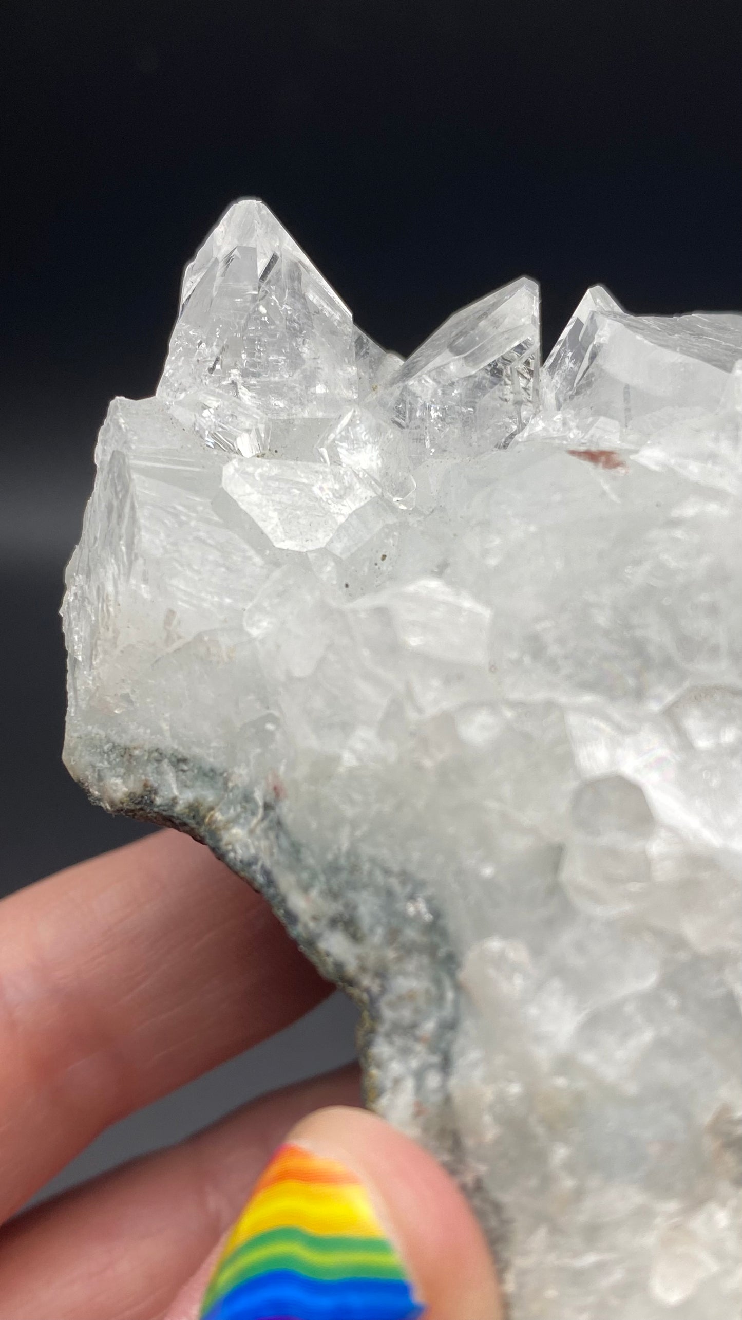 Close-up of a hand holding The Crystalary Apophyllite from the Lonavala Quarry, Pune District, Maharashtra, India. This stunning crystal cluster features several pointed terminations set on a basalt matrix. The person's thumb is visible in the foreground with colorful rainbow-patterned nail design, while the out-of-focus background emphasizes the clarity and apophyllite accents of this beautiful specimen.