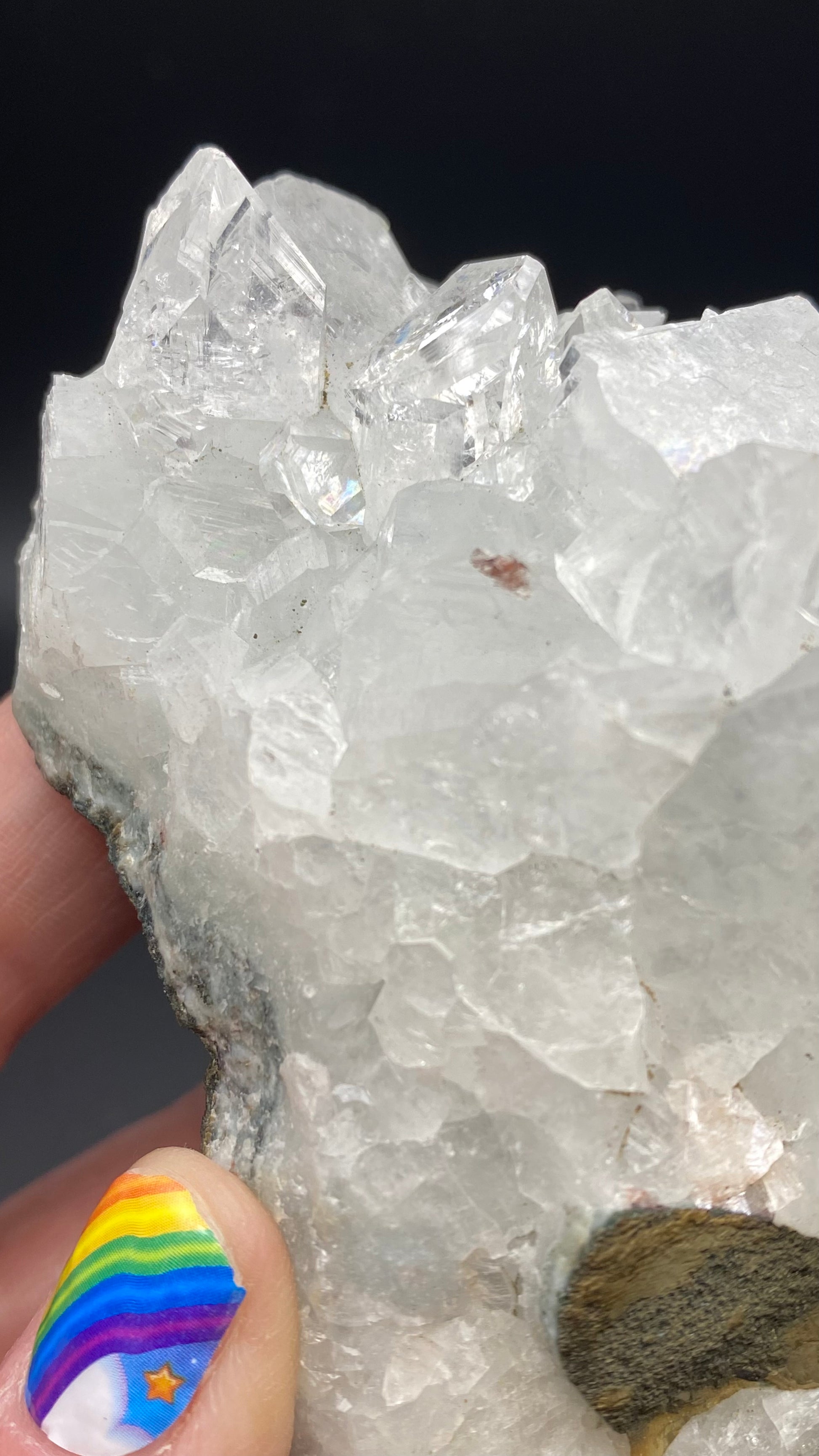 A person with rainbow-themed nail art holds a large, clear Apophyllite crystal cluster from The Crystalary close to the camera. The crystals are jagged and sparkle under the light, featuring a darker mineral inclusion on the lower right side, possibly nestled in a basalt matrix from Lonavala Quarry, Pune District, Pune Division, Maharashtra, India.