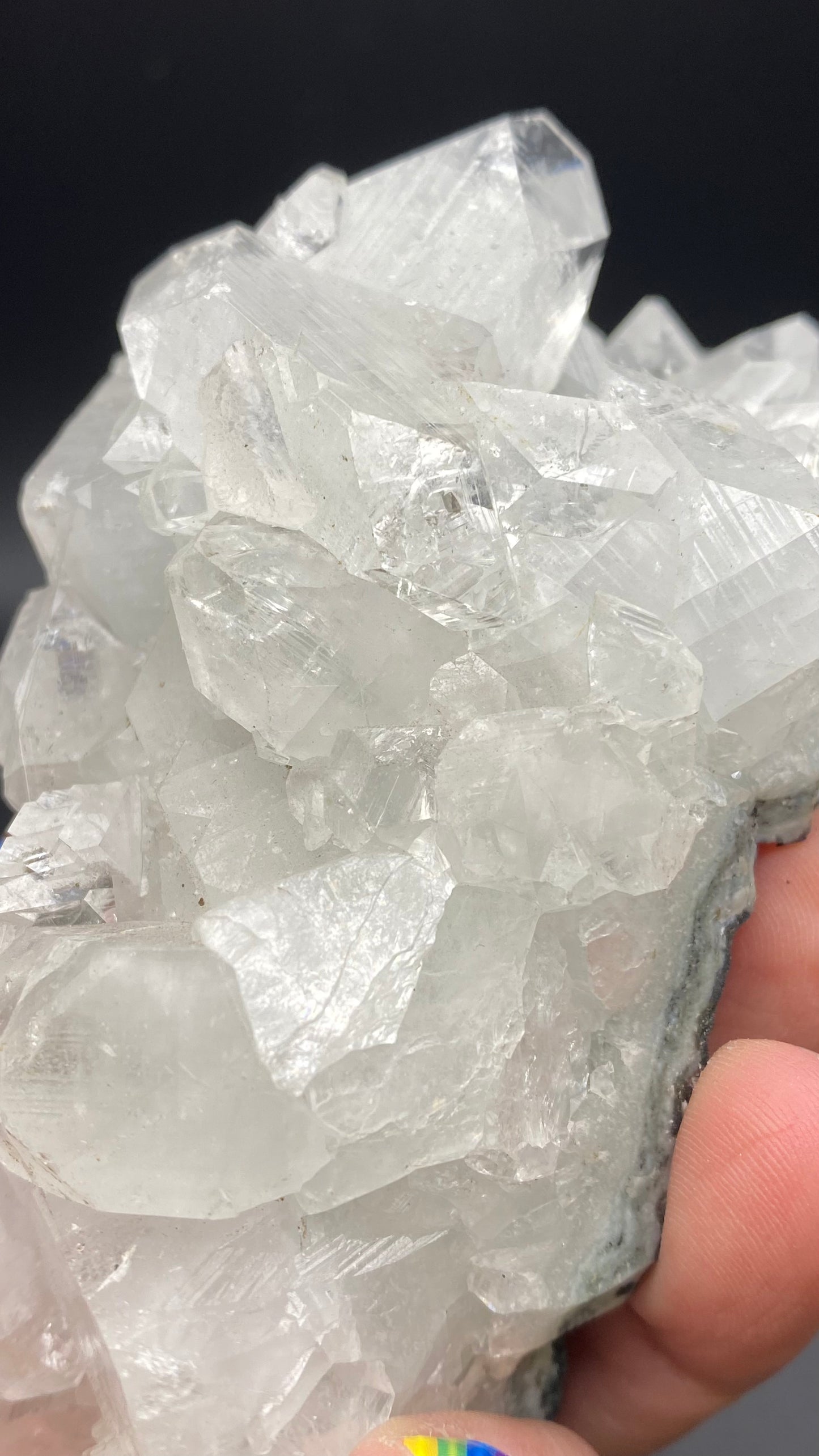 A hand holds a cluster of clear, translucent Apophyllite crystals with sharp, defined edges against a dark background. One fingernail is painted with rainbow-colored stripes, adding a vibrant touch to the mesmerizing display of Apophyllite from the Lonavala Quarry in Pune District, Pune Division, Maharashtra, India. Product by The Crystalary.