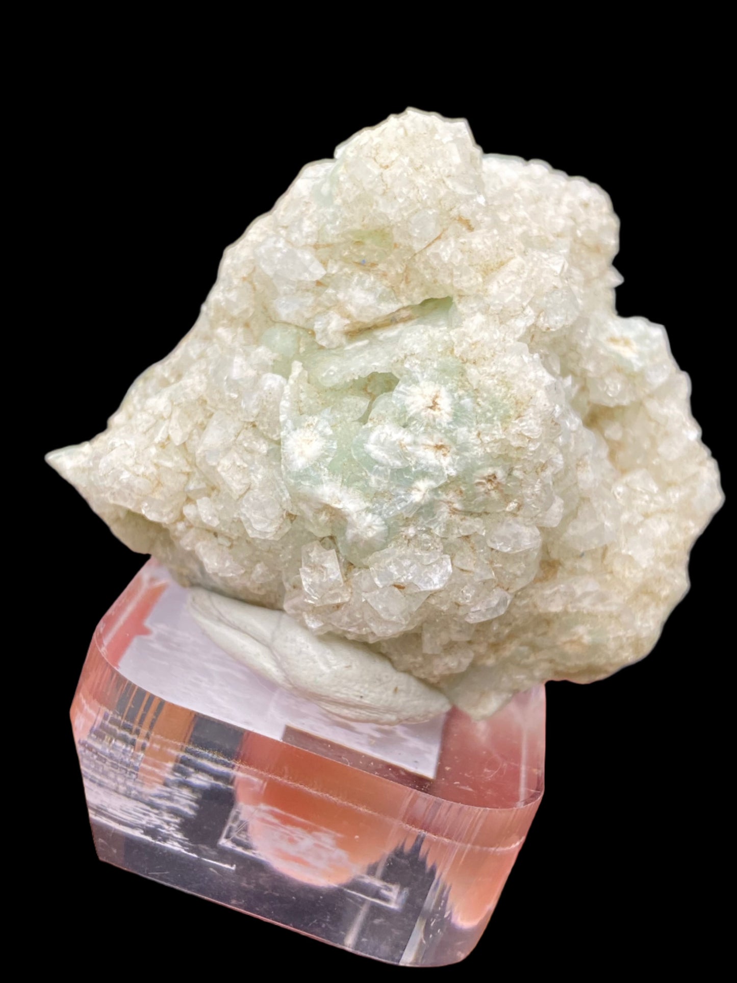 A cluster of translucent, milky white Apophyllite crystal formations rests on a clear and slightly reflective square pedestal. The black background highlights the texture and pale greenish tints within the crystals, which originate from O & G Woodbury Traprock Quarry in Orenaug Hills, Woodbury, Litchfield County, Connecticut. Marketed by The Crystalary, these crystals exhibit a rough and uneven structure.