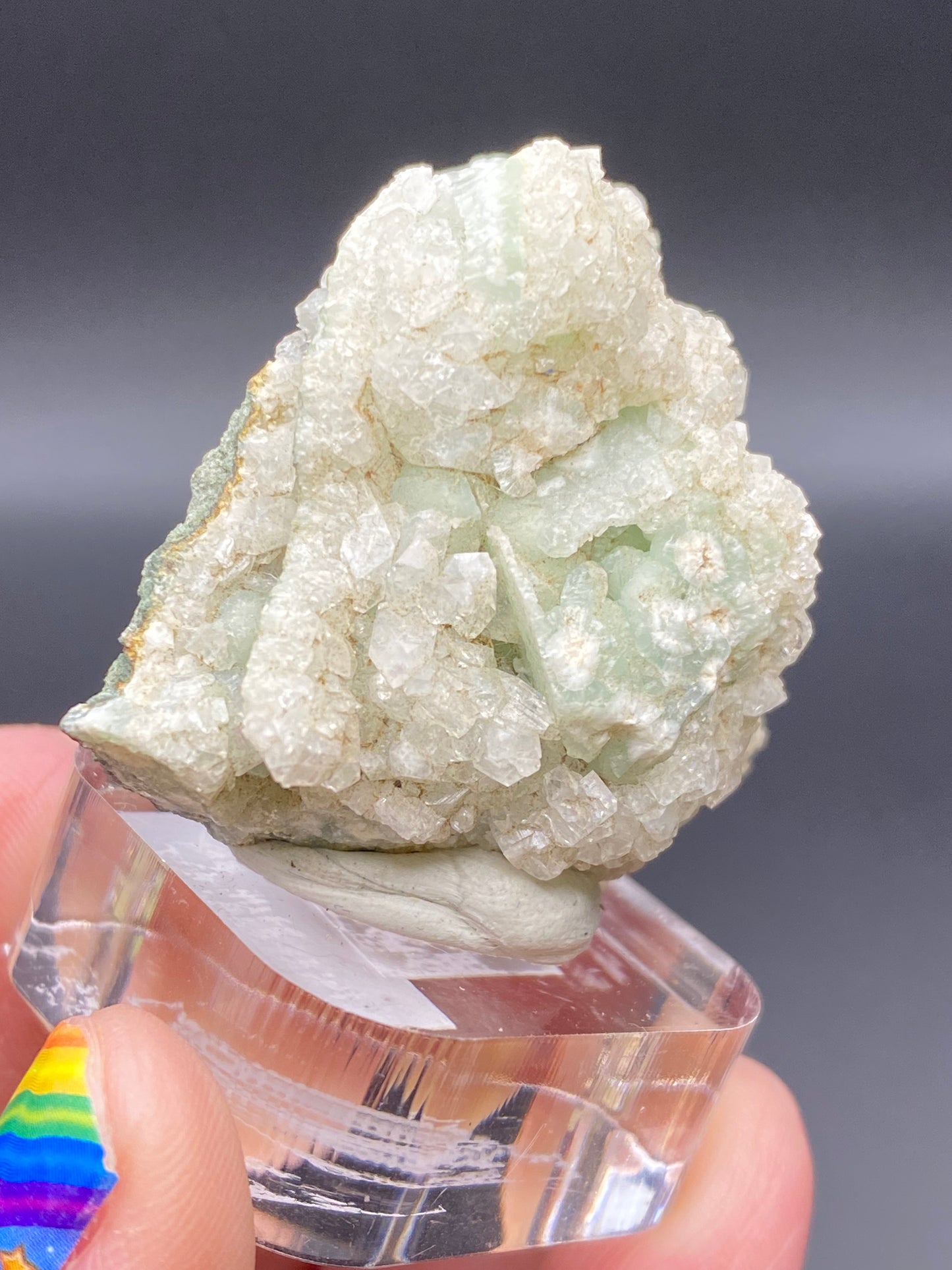 A hand with rainbow-painted nails holds a clear display stand showcasing a stunning crystal formation of green prehnite and white apophyllite, set against a dark, out-of-focus background. This exquisite piece from The Crystalary hails from the renowned O & G Woodbury Traprock Quarry in Orenaug Hills, Litchfield County, Connecticut.