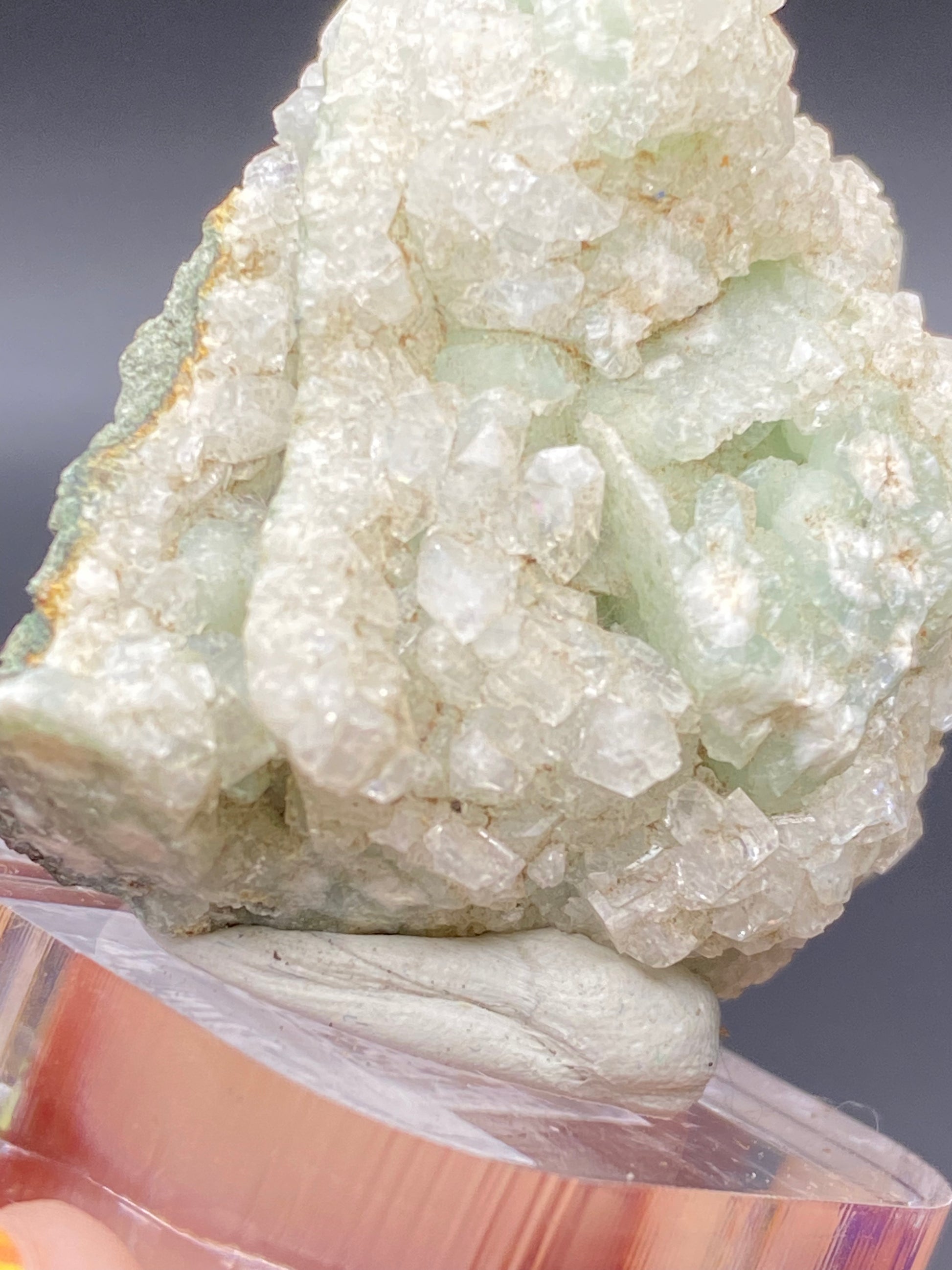 A close-up photograph of a green prehnite mineral specimen with a crystalline structure. The crystals are predominantly light green with some white Apophyllite and transparent formations, displayed on a pinkish-clear stand against a dark background. This stunning piece is named "Apophyllite on Prehnite" and originates from the O & G Woodbury Traprock Quarry in Orenaug Hills, Woodbury, Litchfield County, Connecticut, USA. It is offered by The Crystalary.