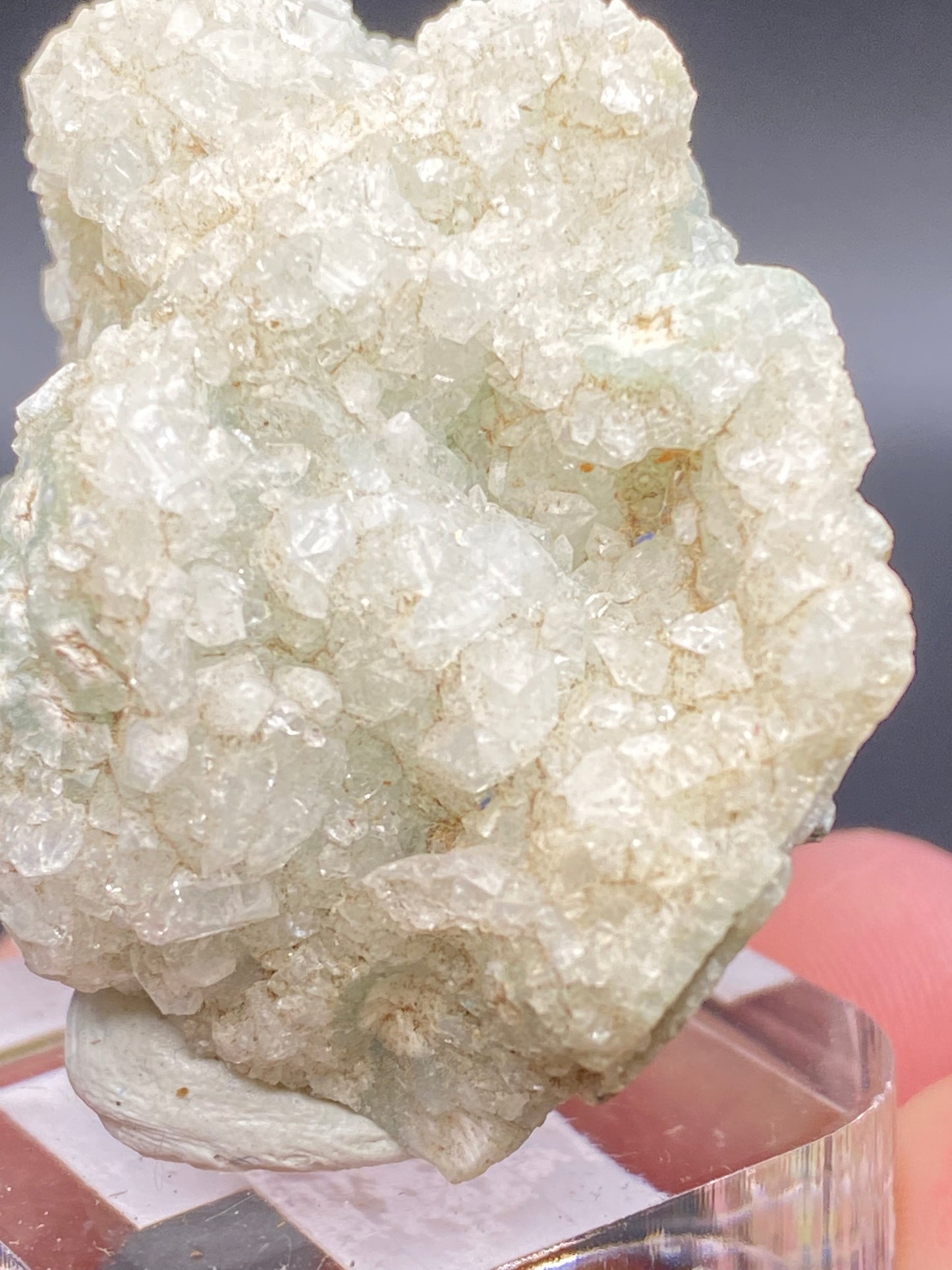 Close-up of a rough, translucent, off-white Apophyllite cluster with green Prehnite accents mounted on a clear display stand. The surface is covered in small, sparkling facets, exhibiting a texture that is both jagged and delicate. A blurred background provides contrast to the crystal's intricate details. This exquisite piece is from O & G Woodbury Traprock Quarry, Orenaug Hills, Woodbury in Litchfield County, Connecticut and offered by The Crystalary.