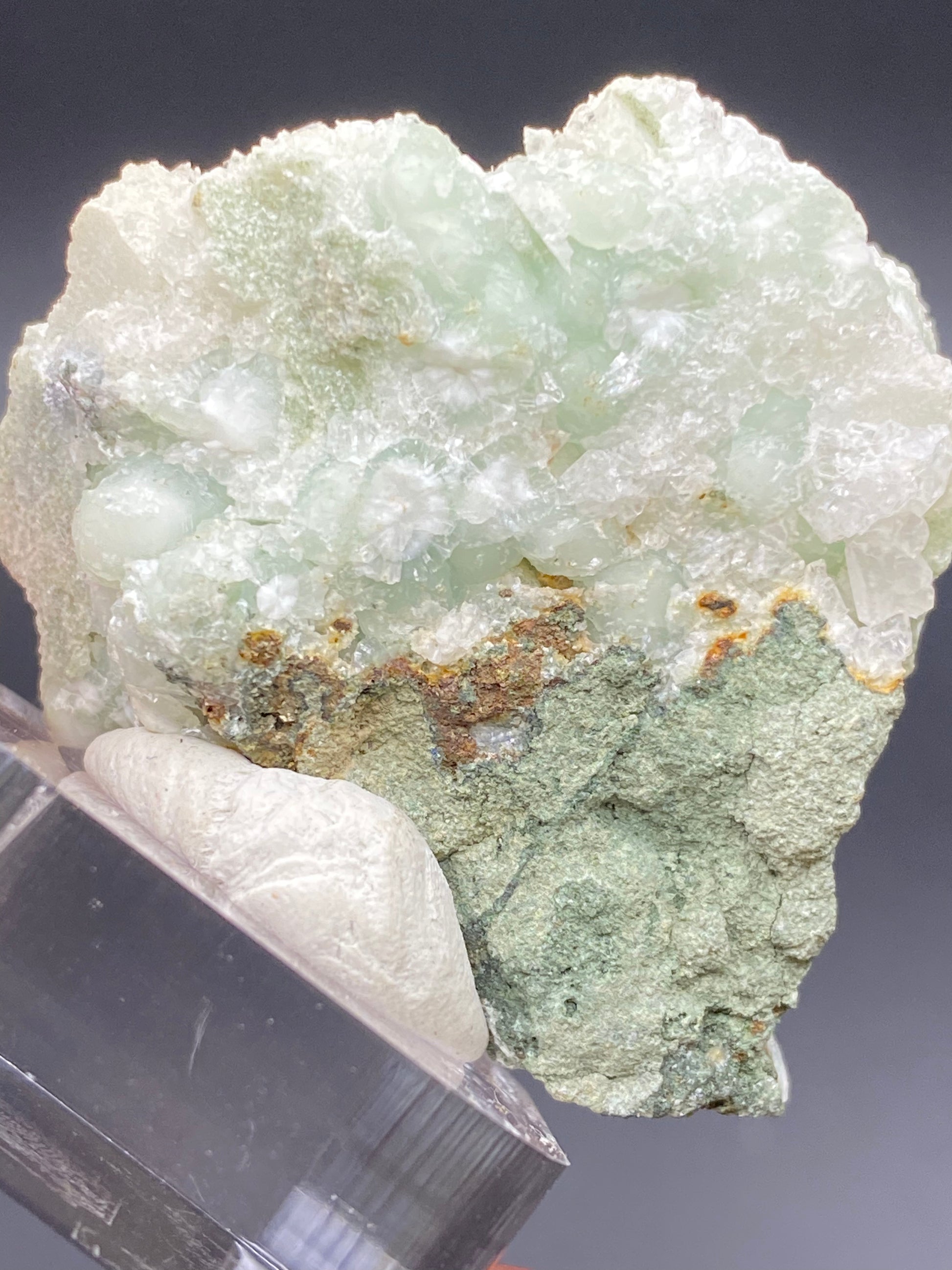 A specimen from The Crystalary named "Apophyllite on Prehnite," sourced from the O & G Woodbury Traprock Quarry in the Orenaug Hills, Woodbury, Connecticut, features a rough, greenish-gray surface with translucent, pale-green prehnite crystalline formations perched atop. Mounted on a clear acrylic stand against a dark background, its uneven and natural texture evokes its origins.