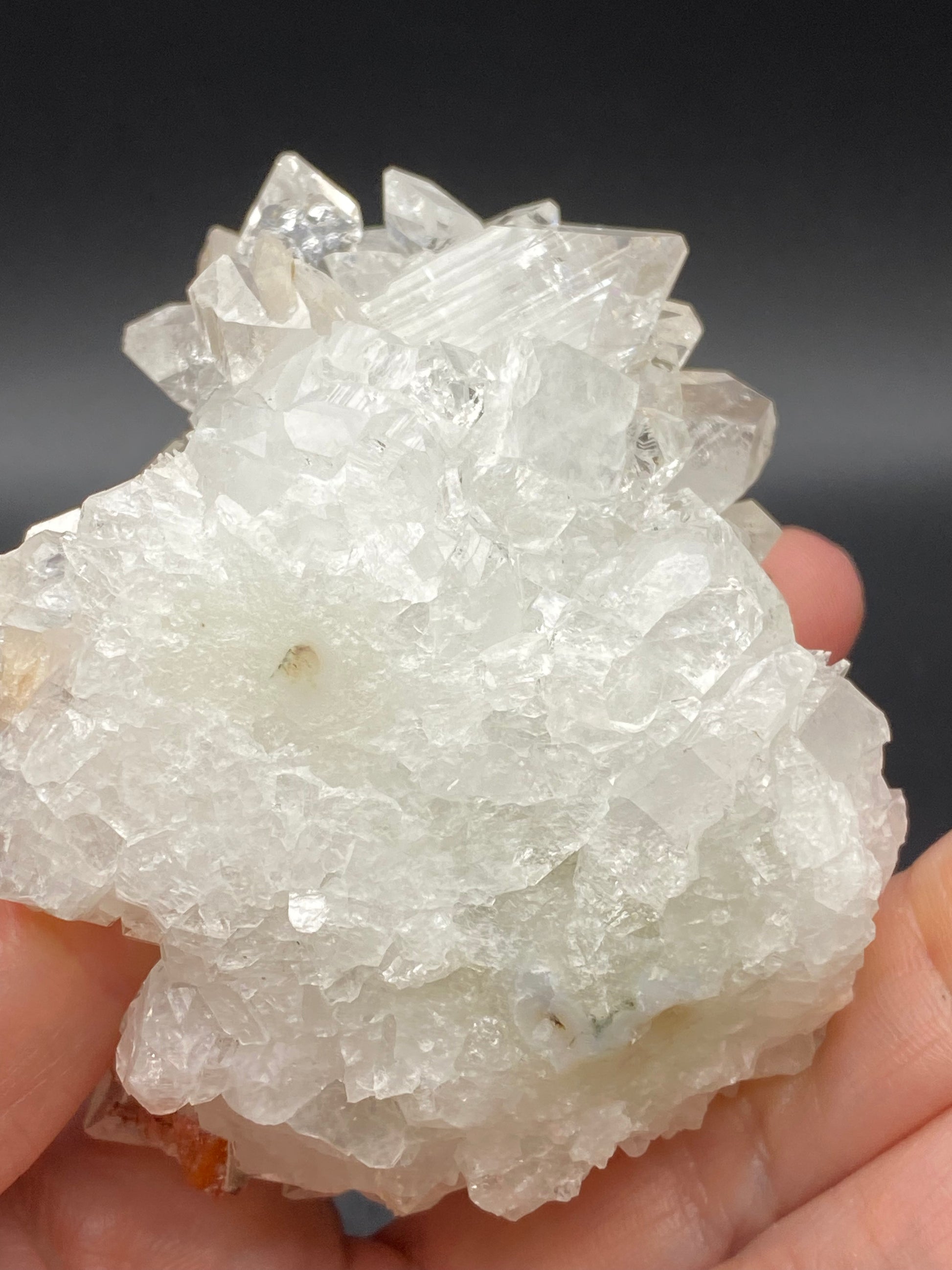 A hand holds a translucent white Apophyllite crystal cluster from The Crystalary against a dark background, showcasing varied formations with some pronounced tips. Amidst the rough and natural appearance of this exquisite mineral from Lonavala Quarry in the Pune District of Maharashtra, India, you can spot Stilbite formations that enhance its intricate geometry.