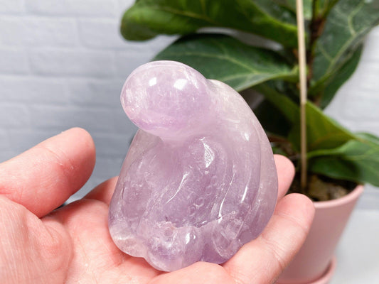 A person is holding a polished, purple Assorted Octopus Crystal Carving from The Crystalary shaped like a sloth in their hand. In the background, there is a green potted plant with broad leaves and a light-colored brick wall, adding to the perfect decorations for planters.