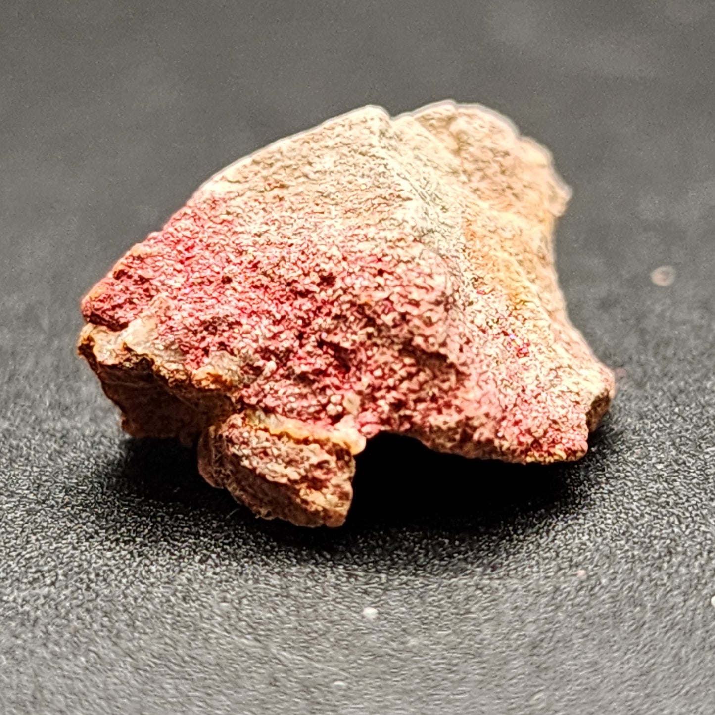 A close-up image features a small, rough piece of rock with a predominantly pinkish hue, tinged with red and some brownish areas. The rock's surface appears textured and granular. This Cinnabar Miniature from The Crystalary is placed on a dark, flat surface and likely originates from the Terlingua District in Brewster County, Texas.