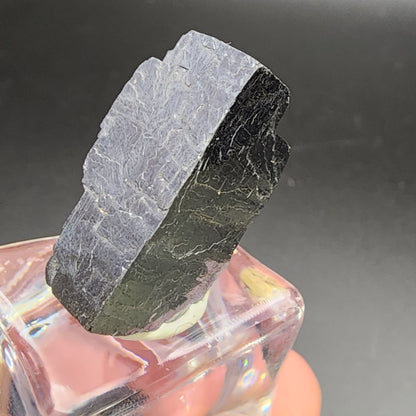 A close-up of a rough, black AUCTION Galena mineral specimen from The Crystalary's collection, sourced from Sweetwater Mine in Ellington, Reynolds County, Missouri, USA. The specimen is displayed on a transparent cubic stand and features an uneven texture with sharp edges. It appears to be slightly tilted against a dark background.