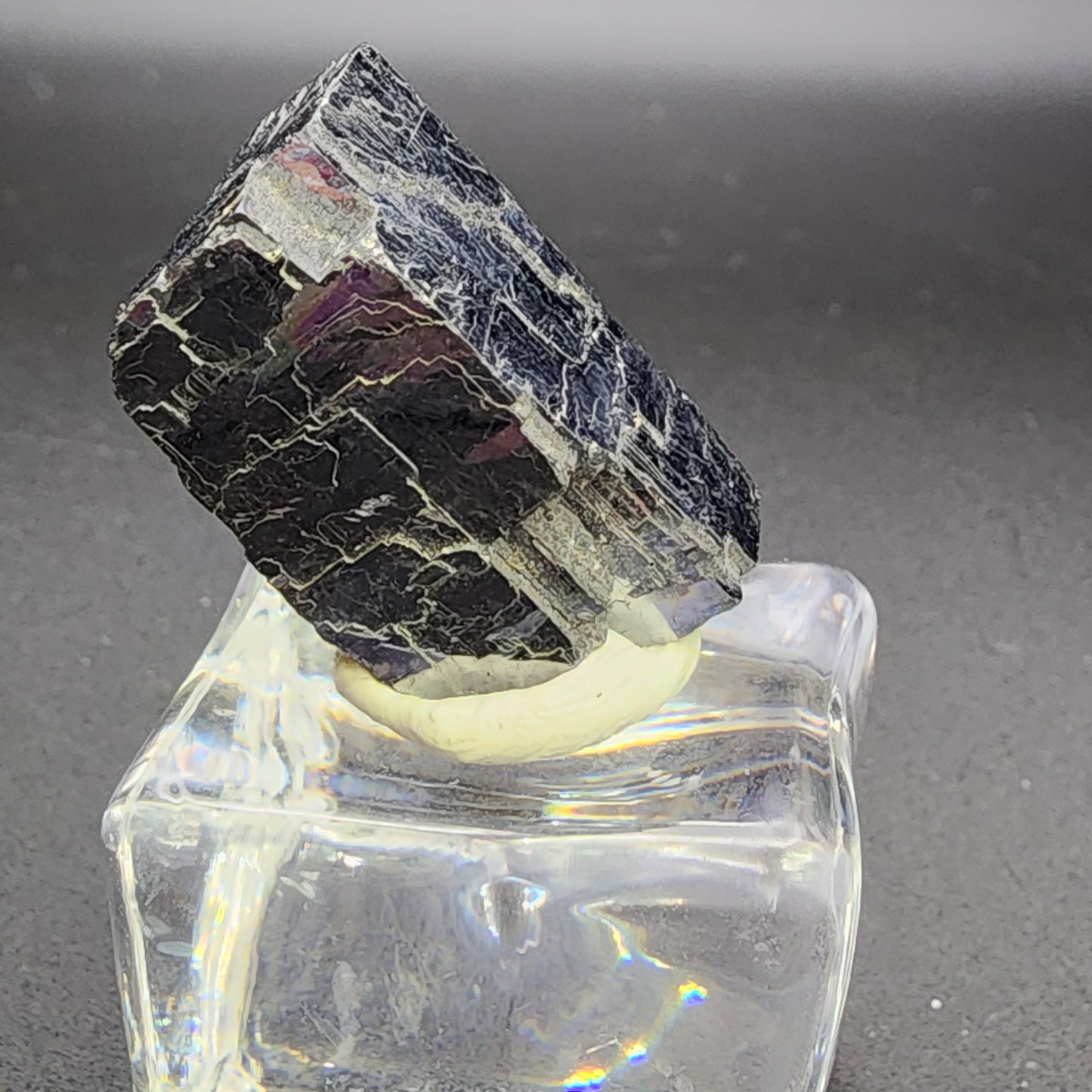 A blocky, dark-colored AUCTION Galena crystal from Sweetwater Mine in Ellington, Reynolds County, Missouri is presented by The Crystalary on a clear, square-shaped base against a simple grey background. The crystal features striations and glossy reflections on its rugged surface that contrast with the smooth, transparent stand.