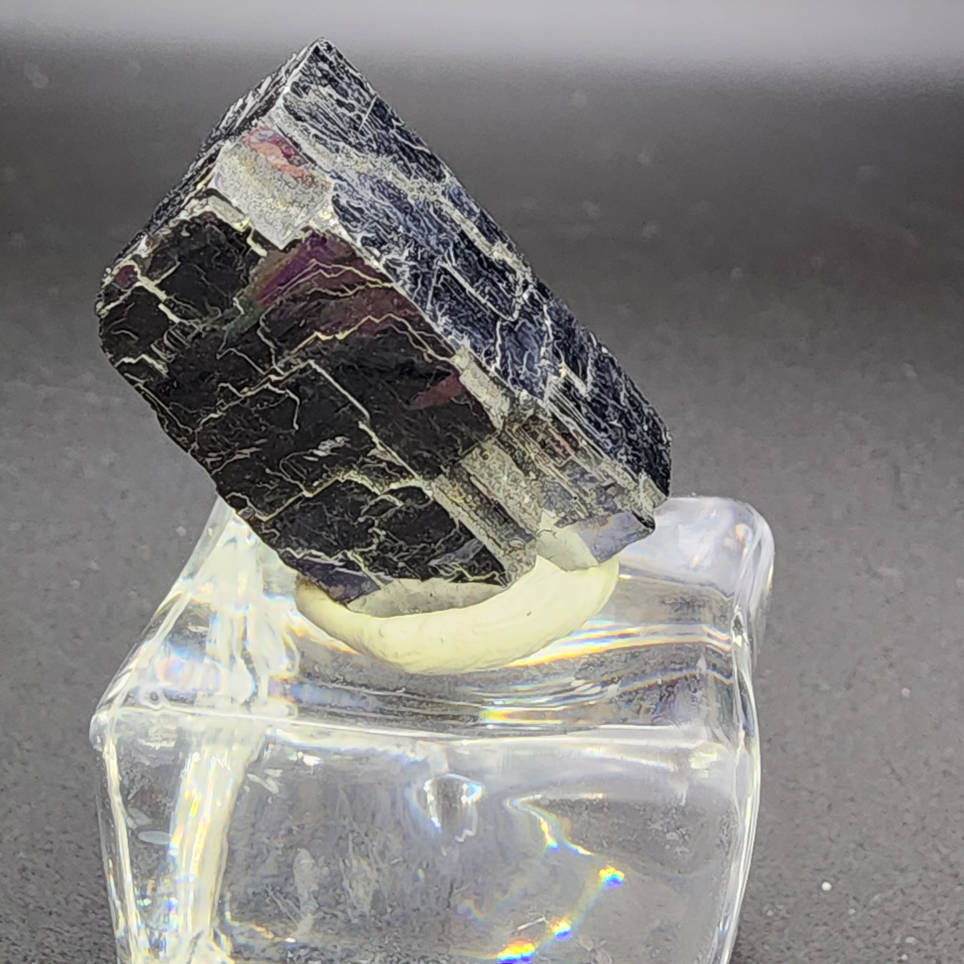 A blocky, dark-colored AUCTION Galena crystal from Sweetwater Mine in Ellington, Reynolds County, Missouri is presented by The Crystalary on a clear, square-shaped base against a simple grey background. The crystal features striations and glossy reflections on its rugged surface that contrast with the smooth, transparent stand.