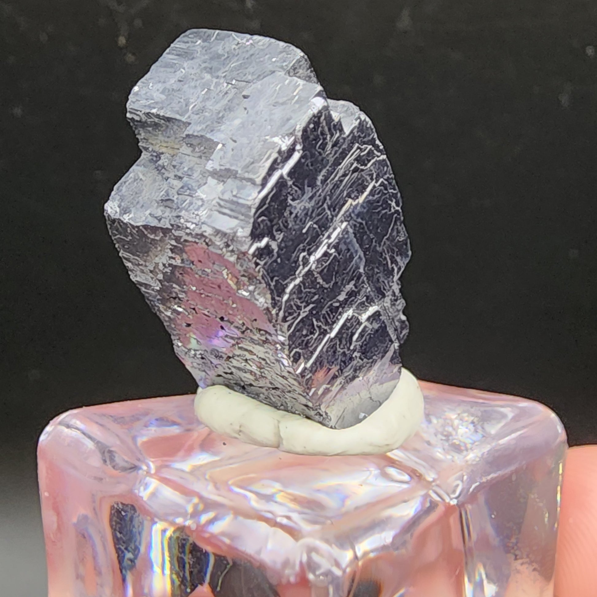 A highly reflective, metallic gray crystal specimen of AUCTION Galena from The Crystalary's Sweetwater Mine in Ellington, Reynolds County, Missouri, USA is mounted on a clear cubic stand with a small amount of light-colored adhesive. The dark background accentuates the shiny, textured surfaces and sharp edges of the crystal.