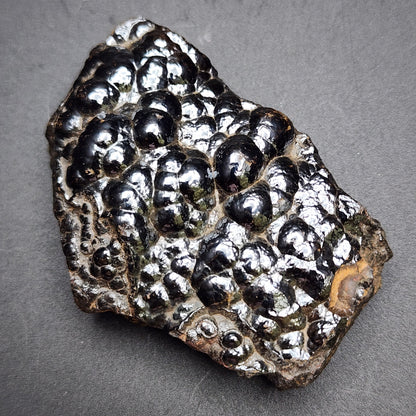 A close-up of a rough specimen of AUCTION- Hematite var. Botryoidal from the Luis Lopez Manganese District, Socorro Co., New Mexico, USA, offered by The Crystalary. The mineral features shiny, bulbous, grape-like formations with a high luster in various shades of black and silver set against a dark, plain background.