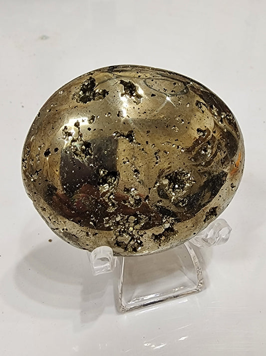 The AUCTION- Pyrite Palm Stone by The Crystalary is showcased on a clear stand against a plain white background. This smooth, polished stone exhibits a metallic gold appearance with small vugs adorned with black inclusions. Its surface reflects light beautifully, emphasizing its shiny and textured features.