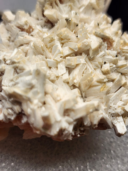 Close-up image of a mineral specimen featuring a dense cluster of pale, elongated crystals, possibly Stilbite and Laumontite from Poona, India. This exquisite piece from The Crystalary’s AUCTION collection showcases tightly packed crystals forming a rough, textured surface. The background is blurred to highlight the detailed structure of the crystals.